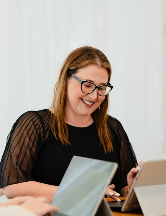 A woman with shoulder-length red hair, glasses, and a black top smiling while looking at a tablet.