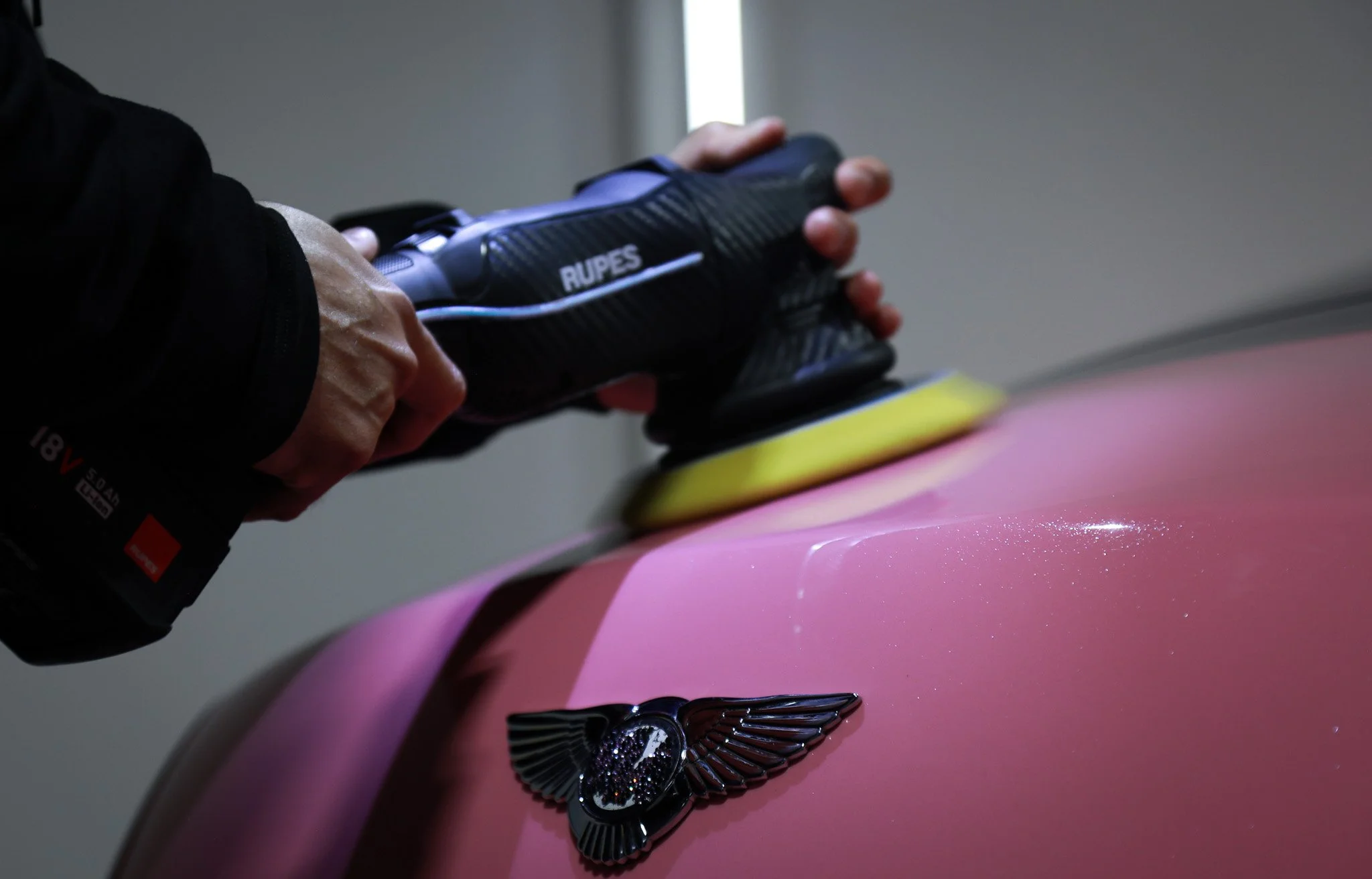 A person polishing a pink car with a black Rupes polisher, showing the car's hood and a black em-flyer emblem.