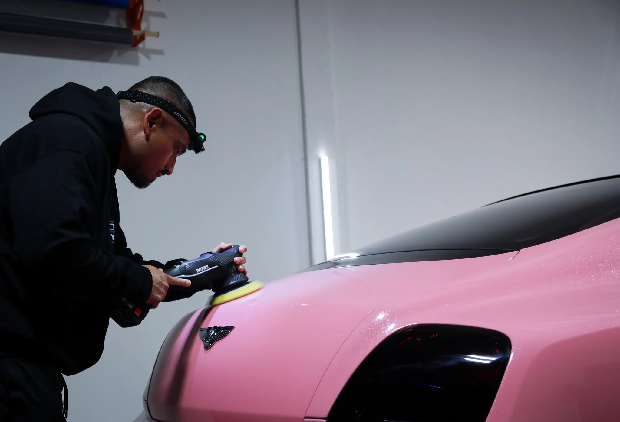 A person wearing a black hoodie and headlamp polishes a pink sports car in a garage.