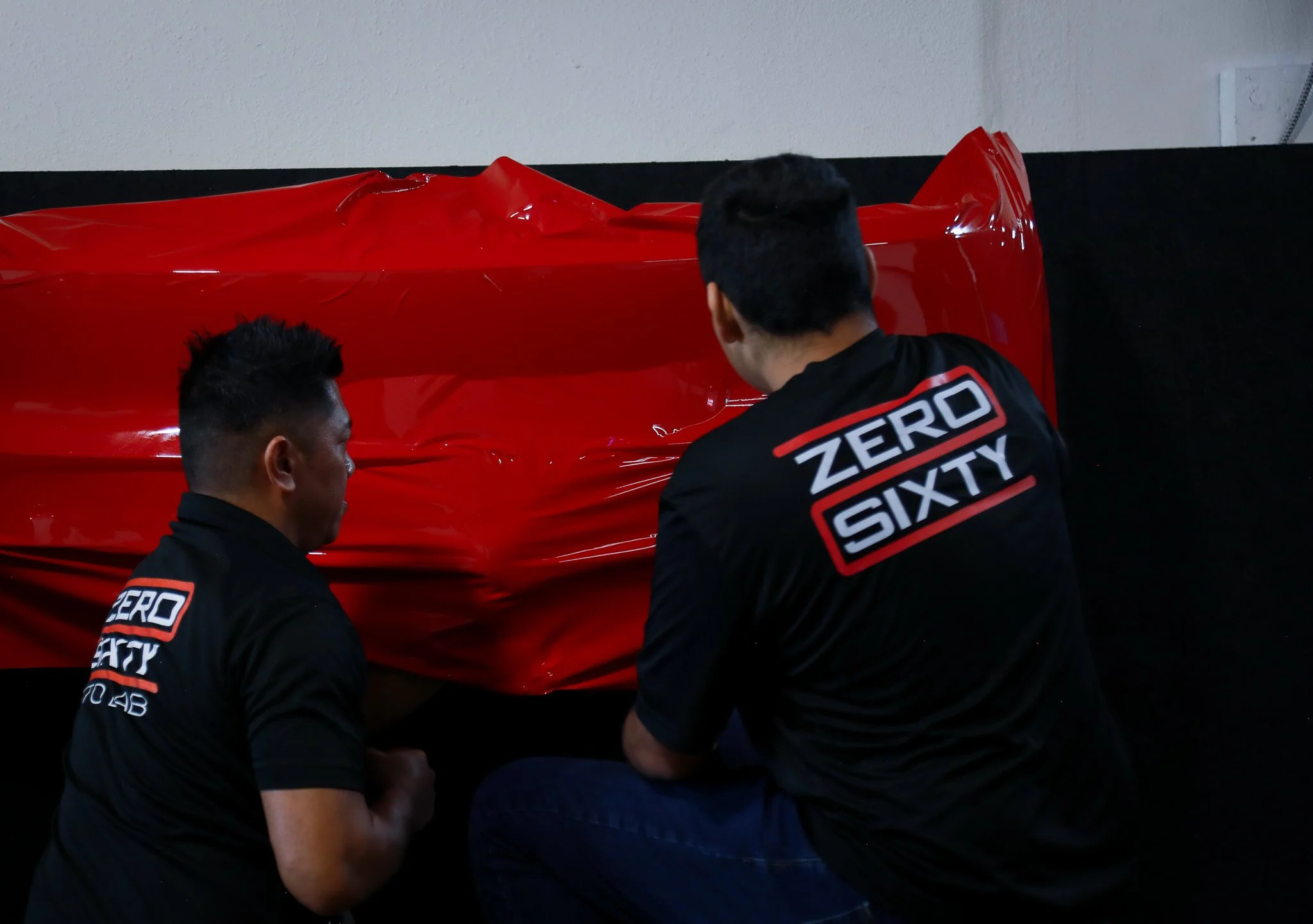 Two men in black shirts with 'Zero Sixty' logos working on a crumpled red car part against a white wall.