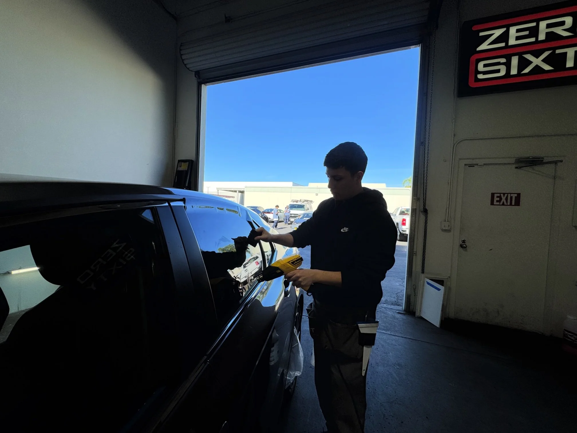 A young man working on a black car inside a garage, using a tool and cleaning the car window. Outside the garage, there is a parking lot with several cars and a blue sky.