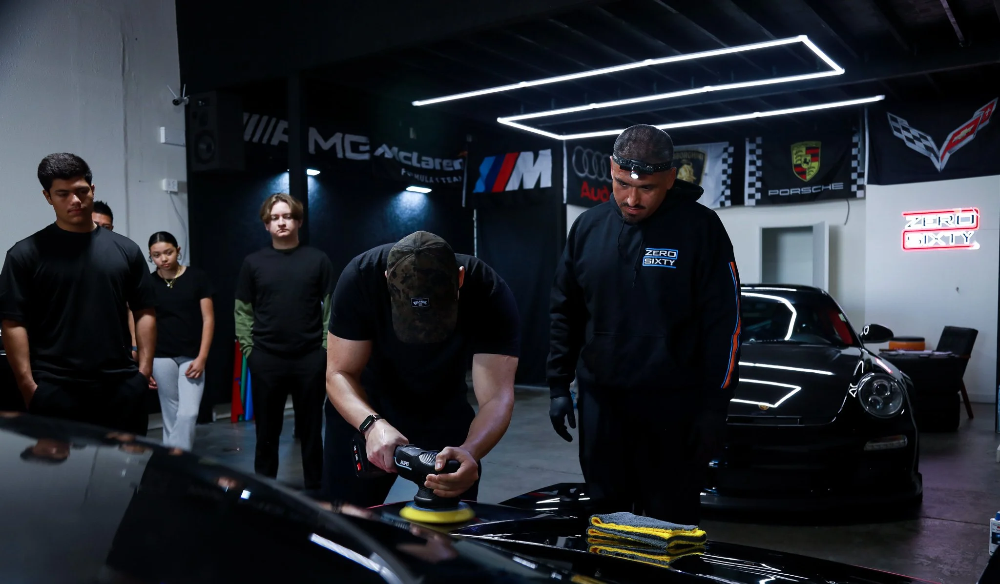 A group of people in a garage with sports cars, with one person polishing a black car using a handheld polishing machine, and others observing, surrounded by automotive banners and neon signs.