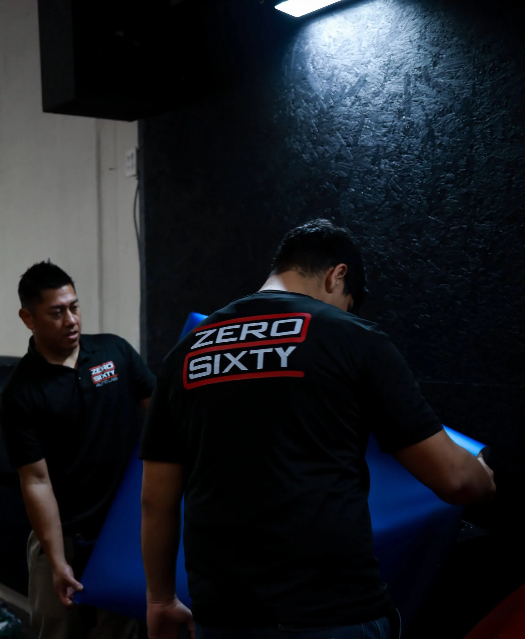 Two men in black shirts with 'ZERO SIXTY' logo handling a blue item in a dimly lit room.