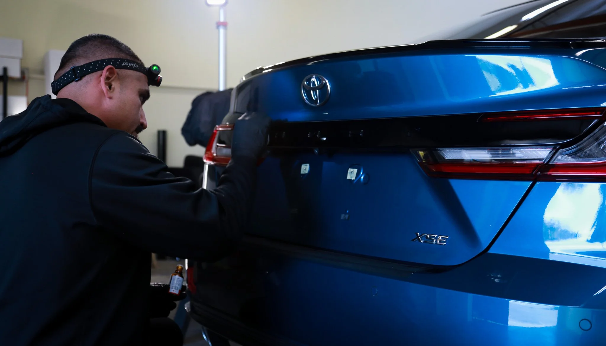 A man wearing a black jacket, black gloves, and a headlamp is inspecting the rear of a blue Toyota XSE car in a garage or workshop.