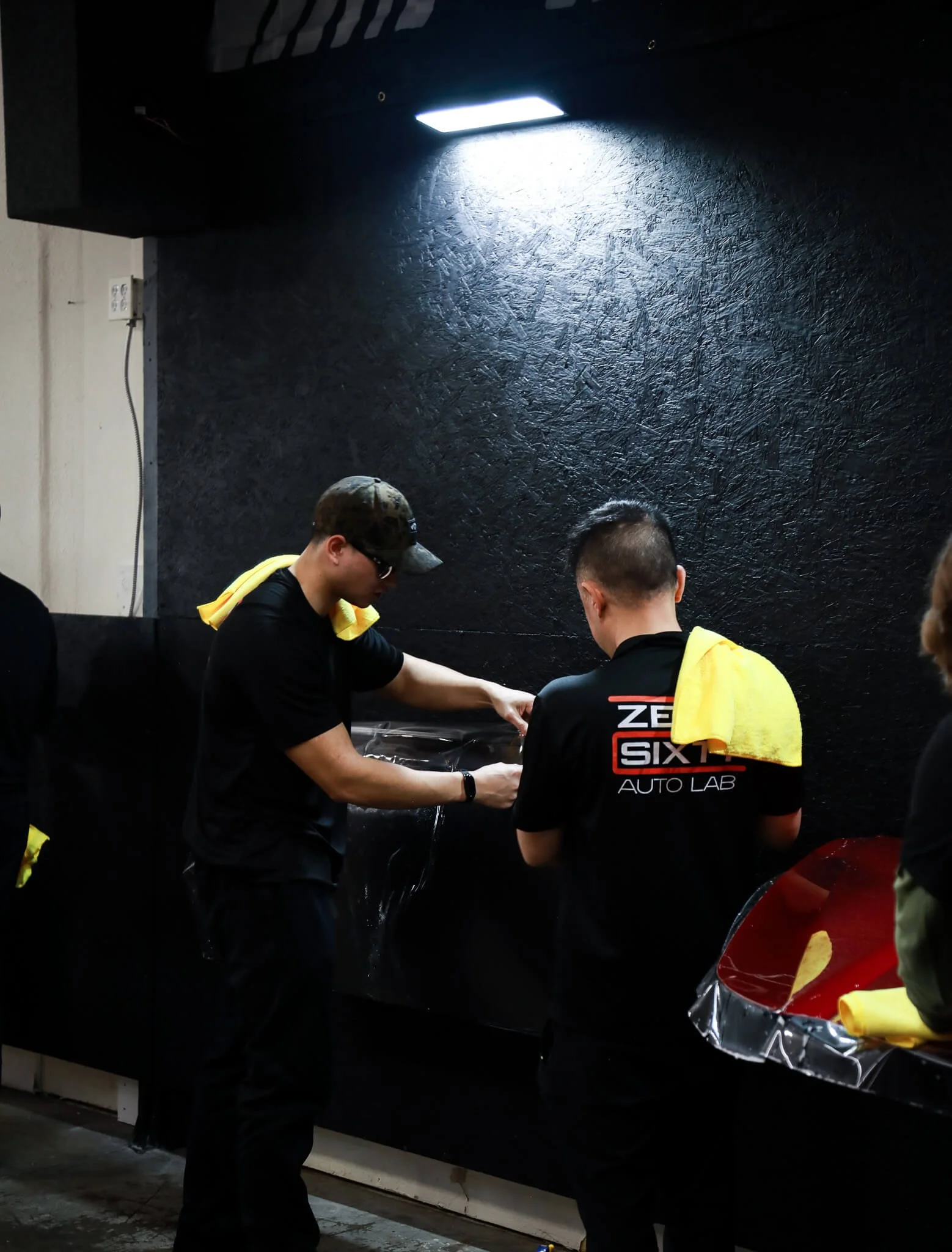 Two men in black shirts with yellow towels on their shoulders stand at a black workbench, working on a car part covered with clear plastic, in a dark workshop with textured black walls and overhead lighting.