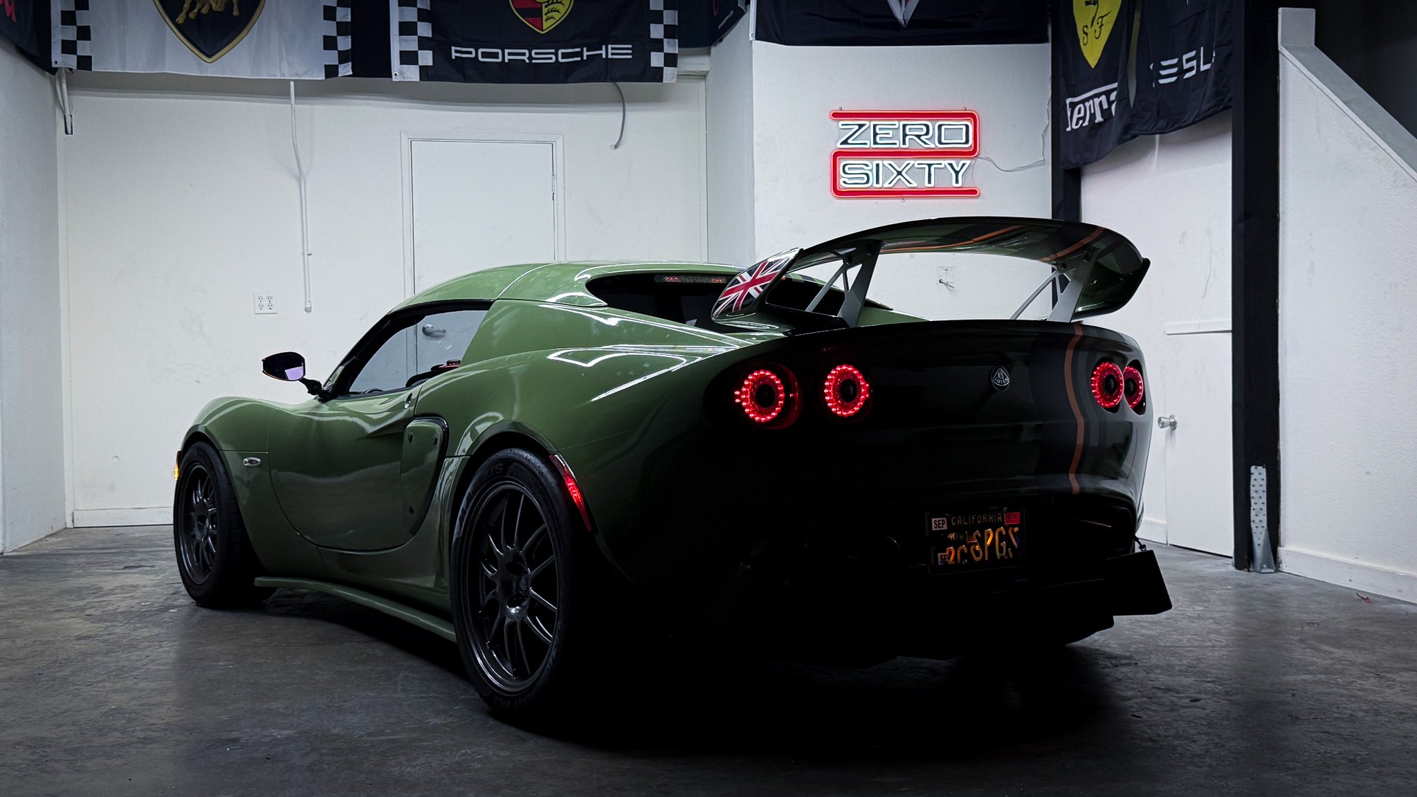 A green Lotus sports car with black rear wing and American license plate in a garage with racing flags and a neon sign that says 'Zero Sixty' in red and white.