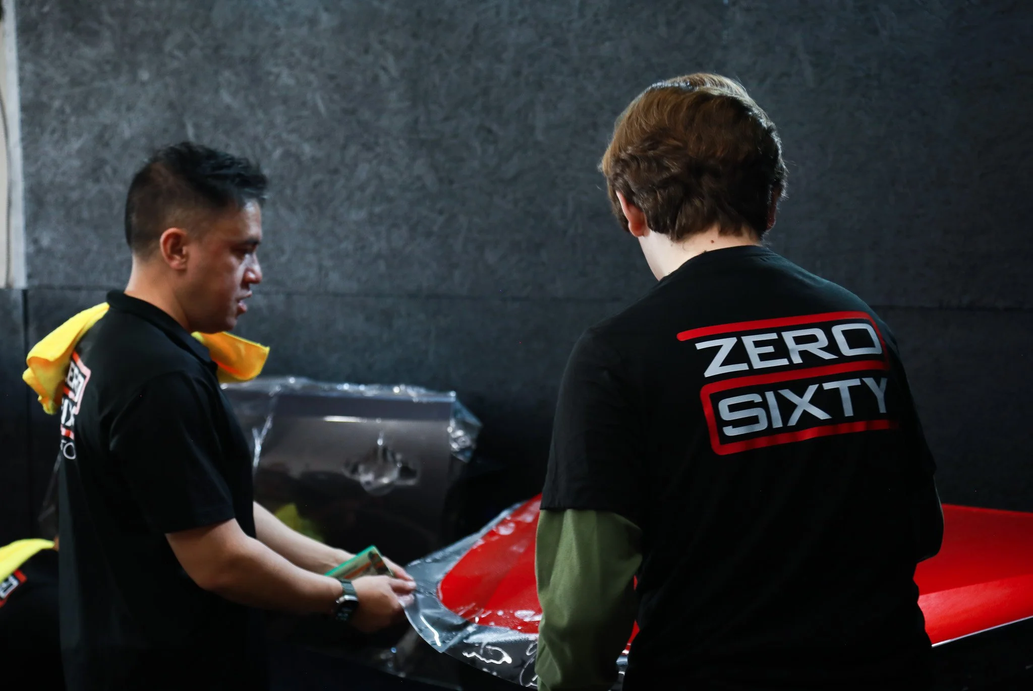Two men examining a red car part, one is wearing a Black shirt with yellow accents and the other a Black shirt with 'ZERO SIXTY' embroidered on the back.