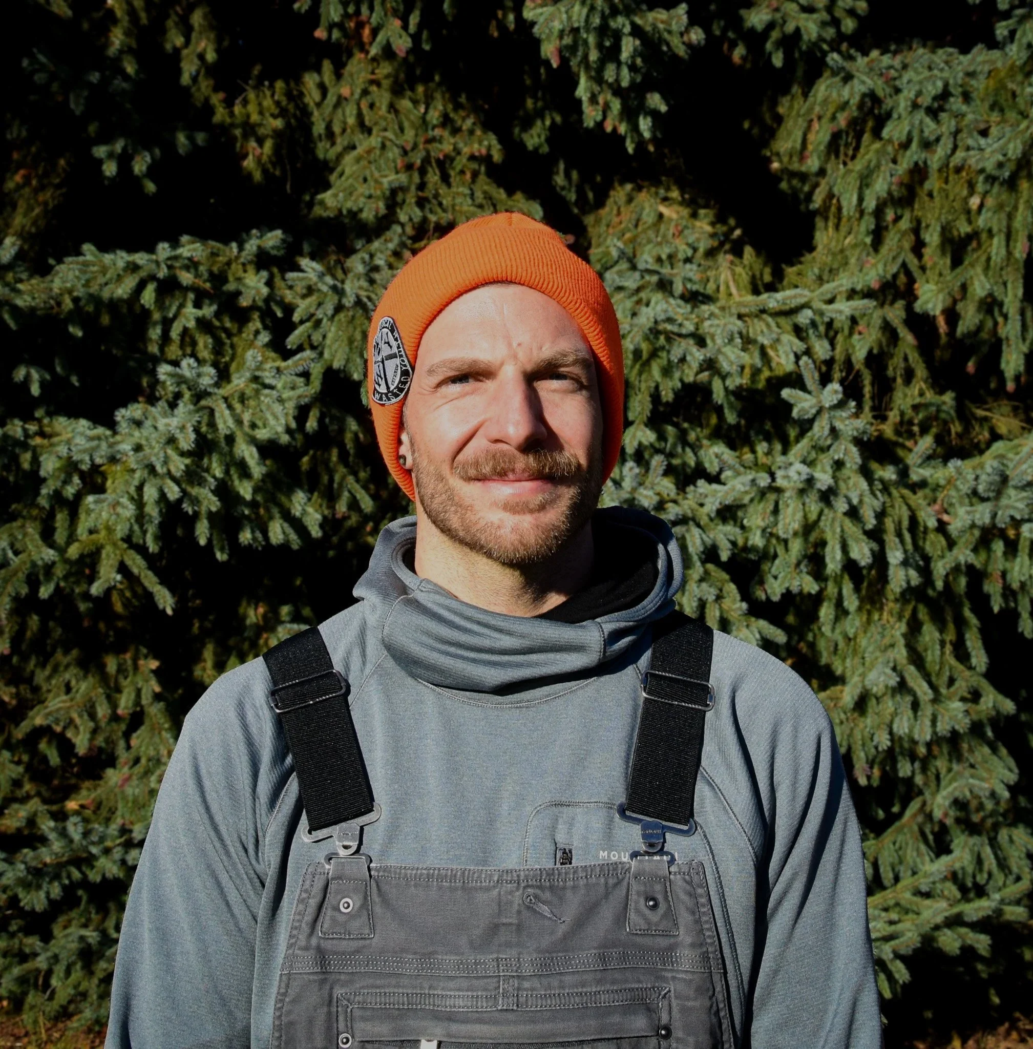Murray Marshal. An electrician outdoors in front of evergreen trees, wearing an orange beanie, gray jacket, and black overalls, sunlight on his face.