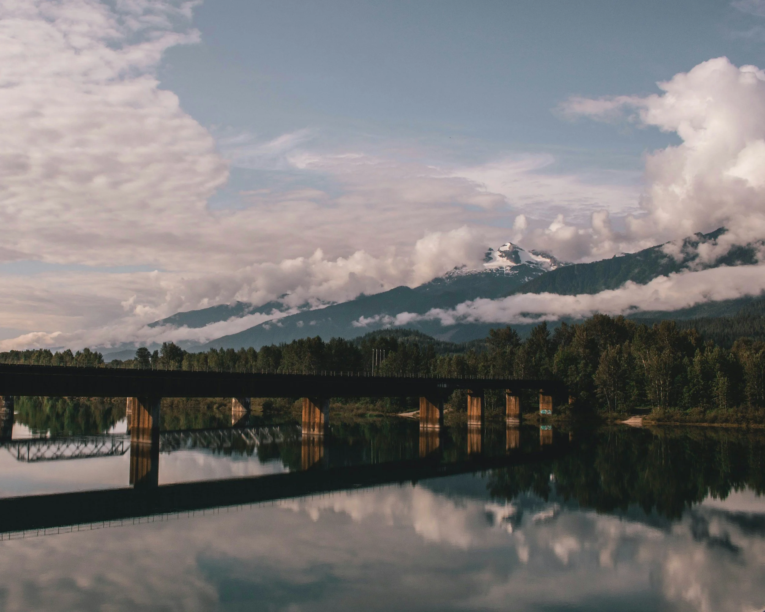 A landscape with a bridge over a calm body of water, reflecting a mountain with snow on its peak surrounded by trees under a cloudy sky.