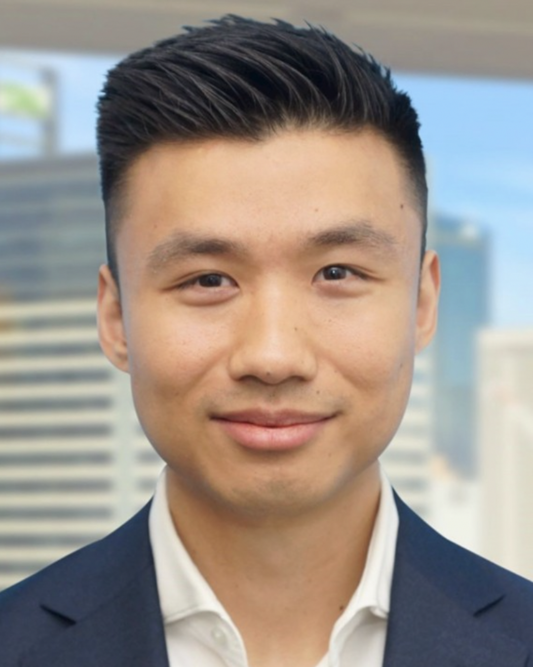 Close-up of a young man with short black hair, wearing a dark suit jacket and a white shirt, standing outdoors with a city skyline in the background.