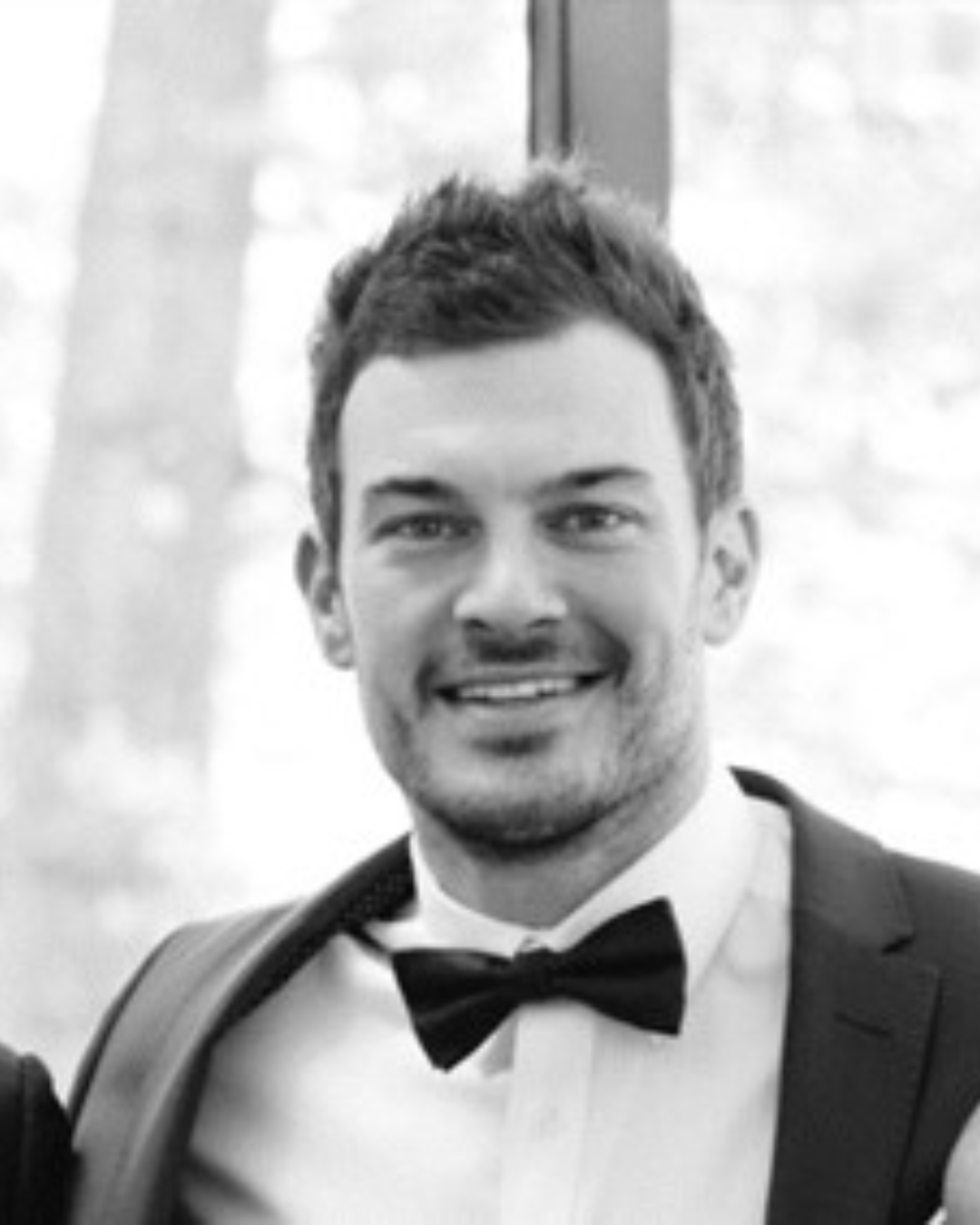 Black and white photo of a smiling man wearing a tuxedo and bow tie, outdoors with blurred trees in the background.