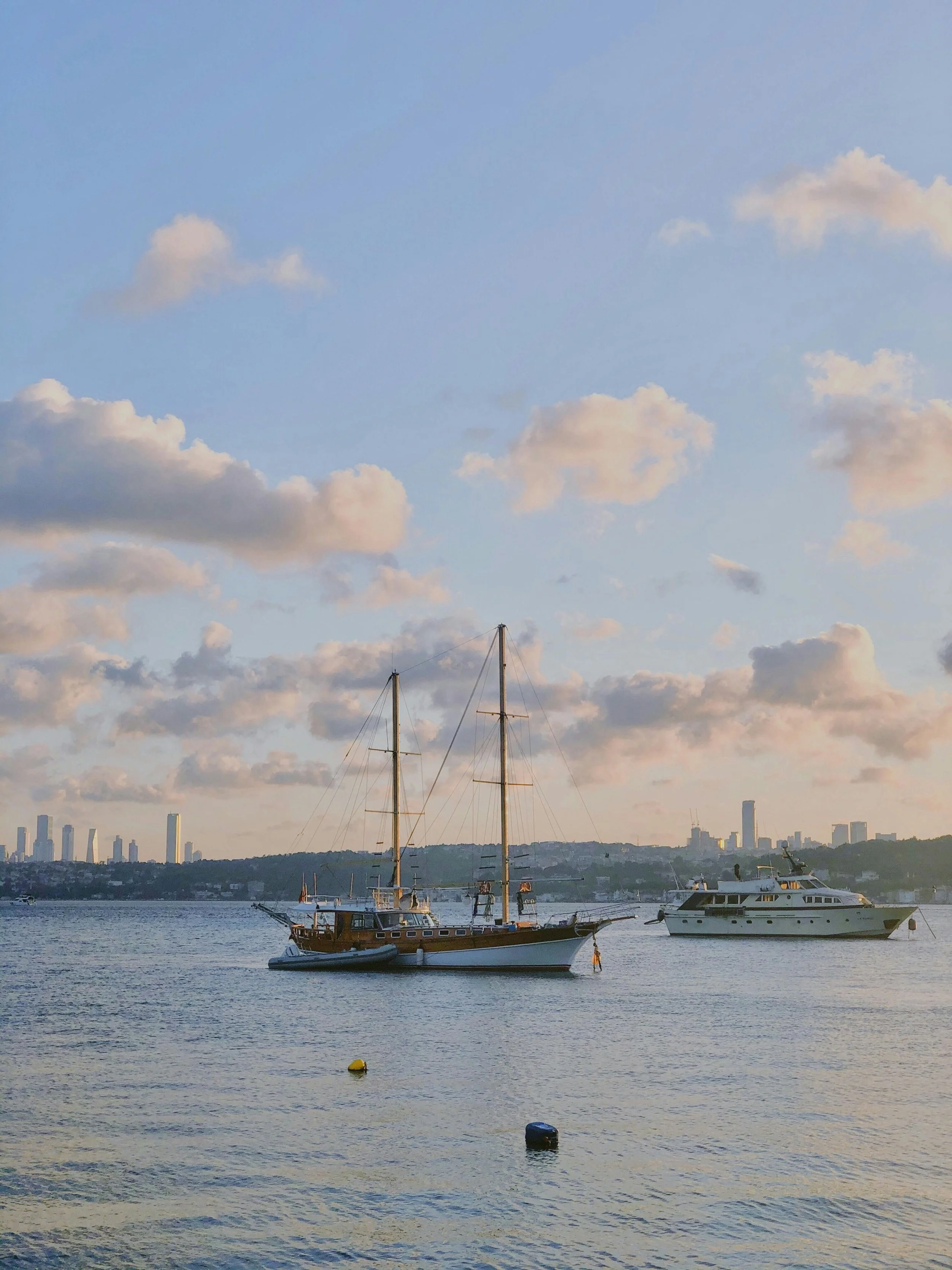 Two boats, a sailboat and a yacht, floating on a calm body of water with city skyline in the distance and a partly cloudy sky above