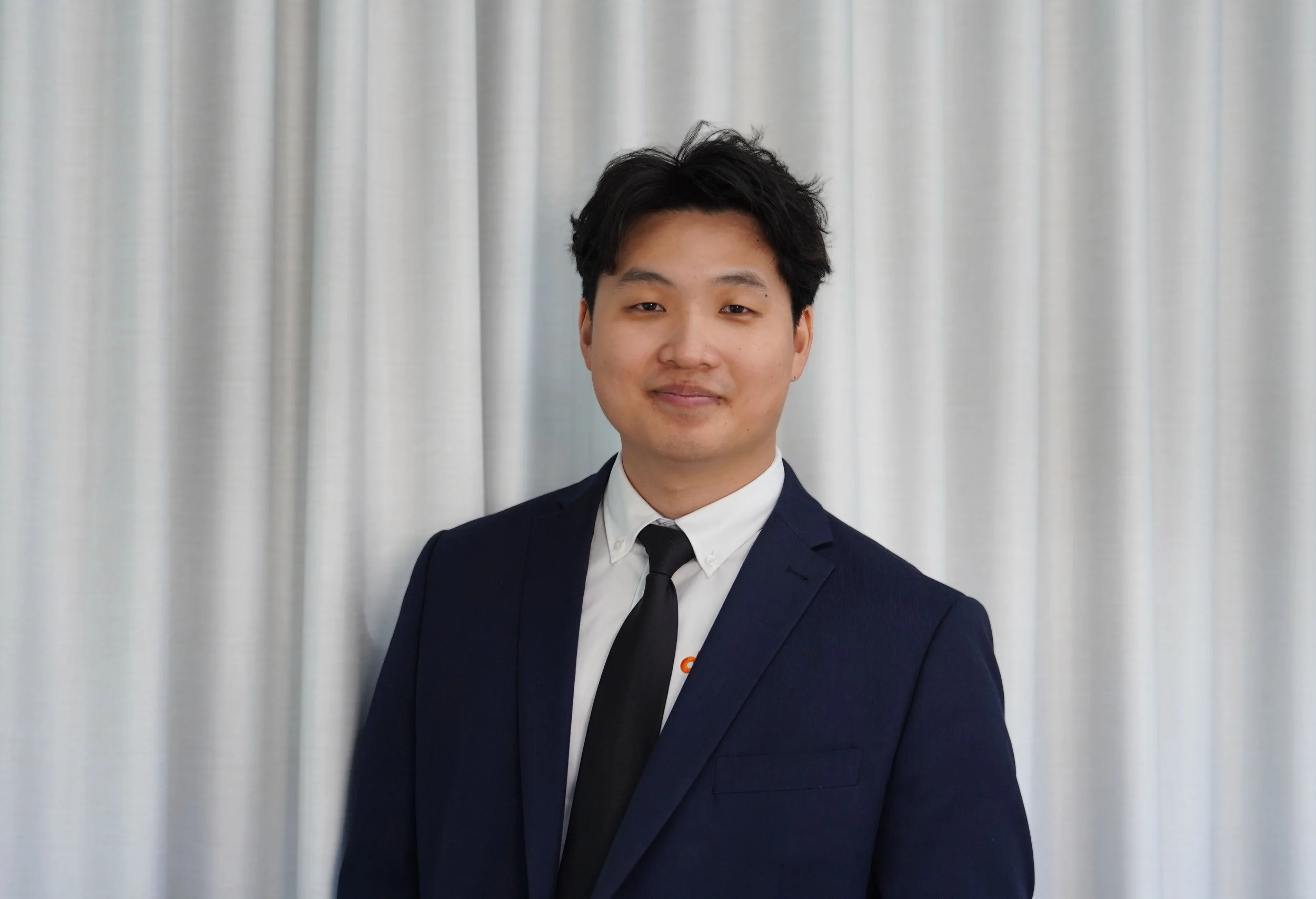 A young man in a navy blue suit with a white shirt and black tie standing in front of a light-colored curtain.