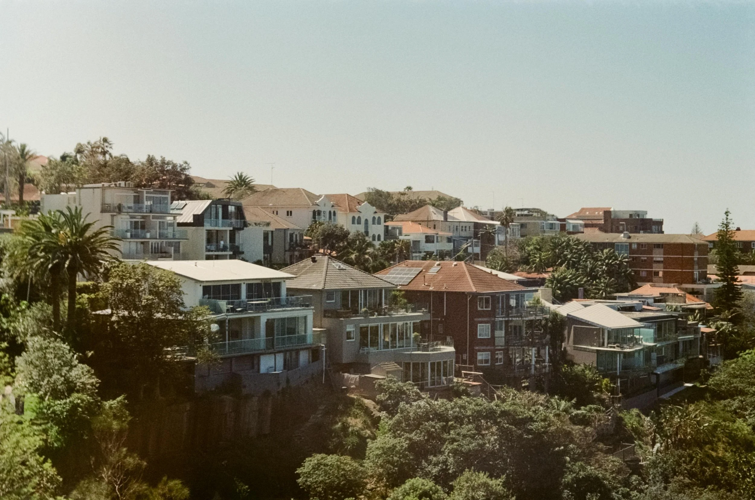 Residential houses in Australia with various roof styles and colors on a hillside, surrounded by greenery and trees, under a clear sky.