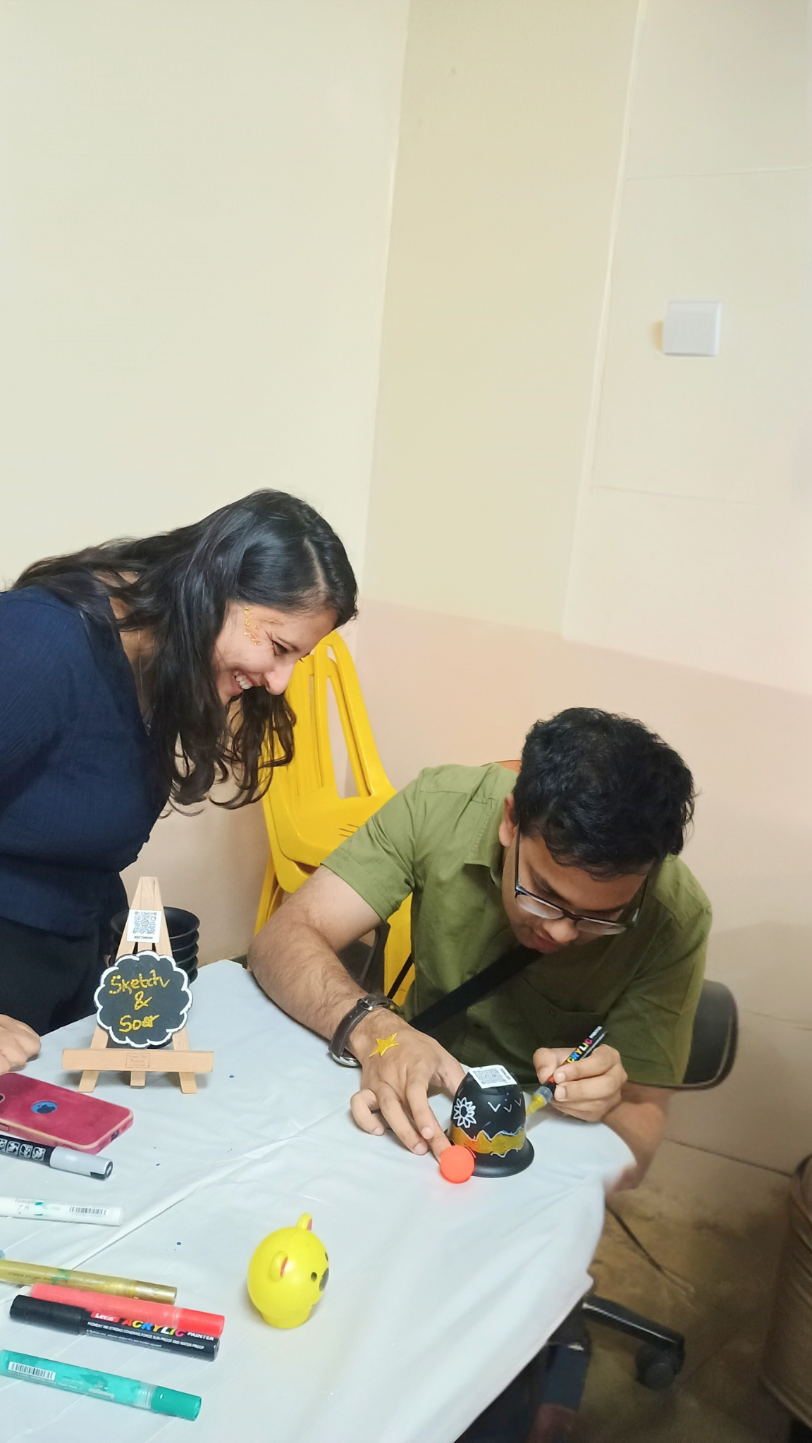 Two people decorating a black round object at a table with various markers and craft supplies, one smiling while the other is focused on coloring
