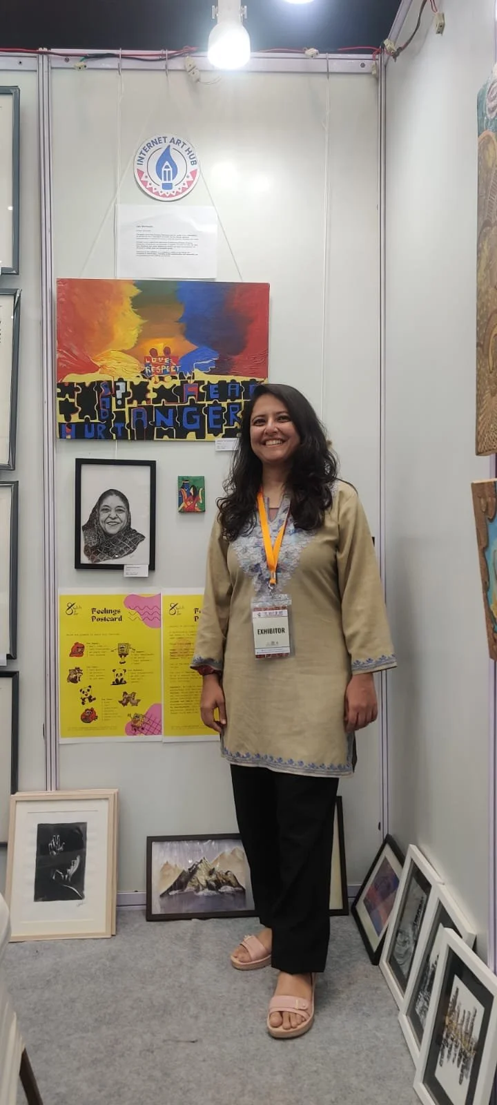 A woman smiling at an art exhibition booth with artwork and posters behind her.