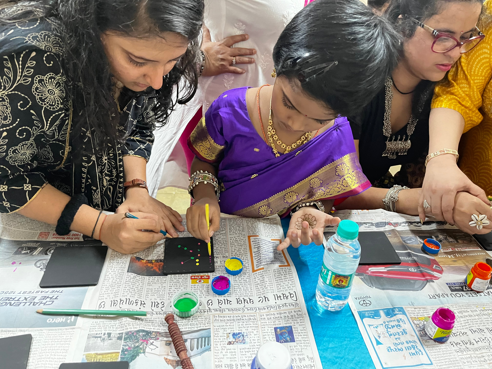 Women decorating small black objects with colorful dots using paint in an indoor setting, with newspapers on the table and various art supplies around.