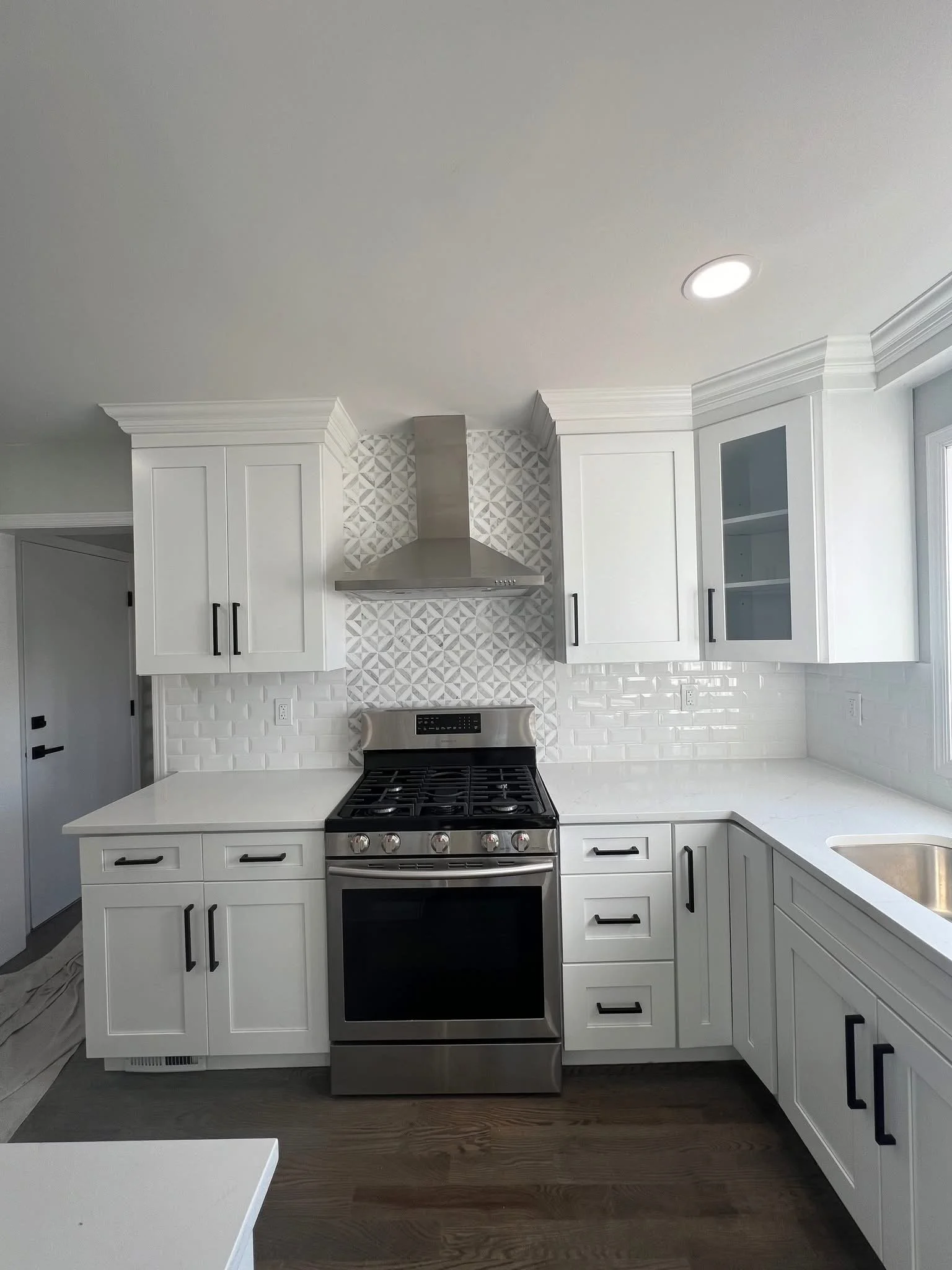 White kitchen with stainless steel stove, white cabinetry with black handles, a patterned backsplash, and a sink near a window.