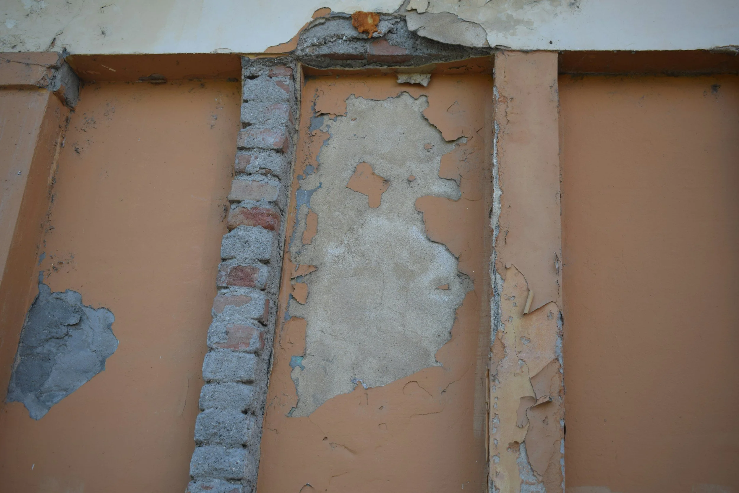 A close-up of a wall with peeling paint, exposing layers of plaster and brickwork underneath.