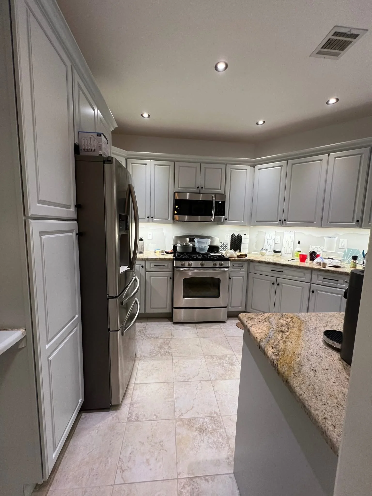 Modern laundry room with white cabinets, stainless steel refrigerator, microwave, and oven, beige tiled floor, and cluttered countertop with various items.