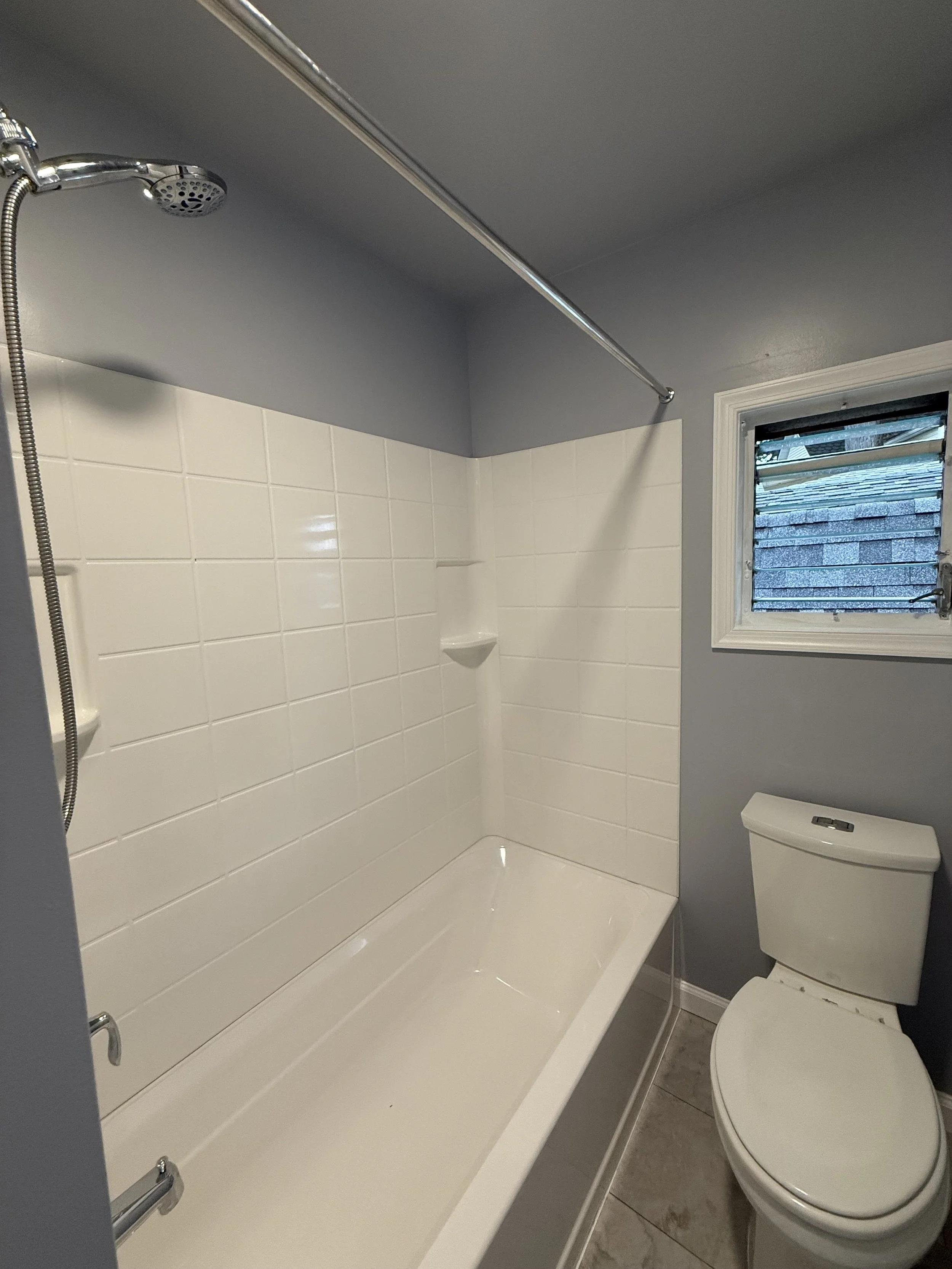 Small bathroom with a shower-tub combination, white tiled wall, gray walls, a small window with louvers, and a white toilet.