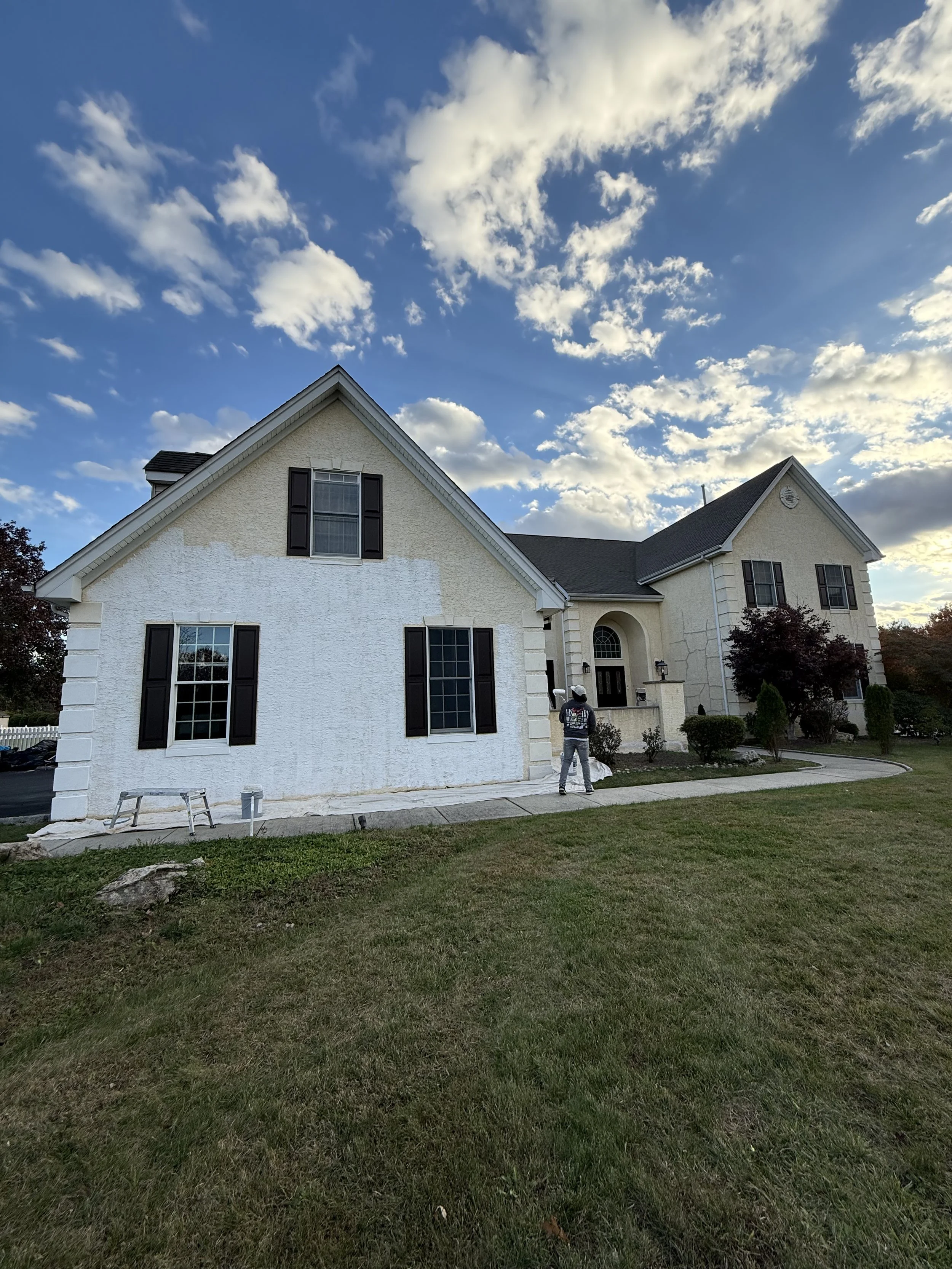 A house undergoing exterior painting with one house wall painted white and the upper part unpainted, three windows with black shutters, a front entryway with an arched window above the door, a man in a grey hoodie and cap standing outside, well-maintained lawn with bushes and trees, and a partly cloudy sky at sunset.