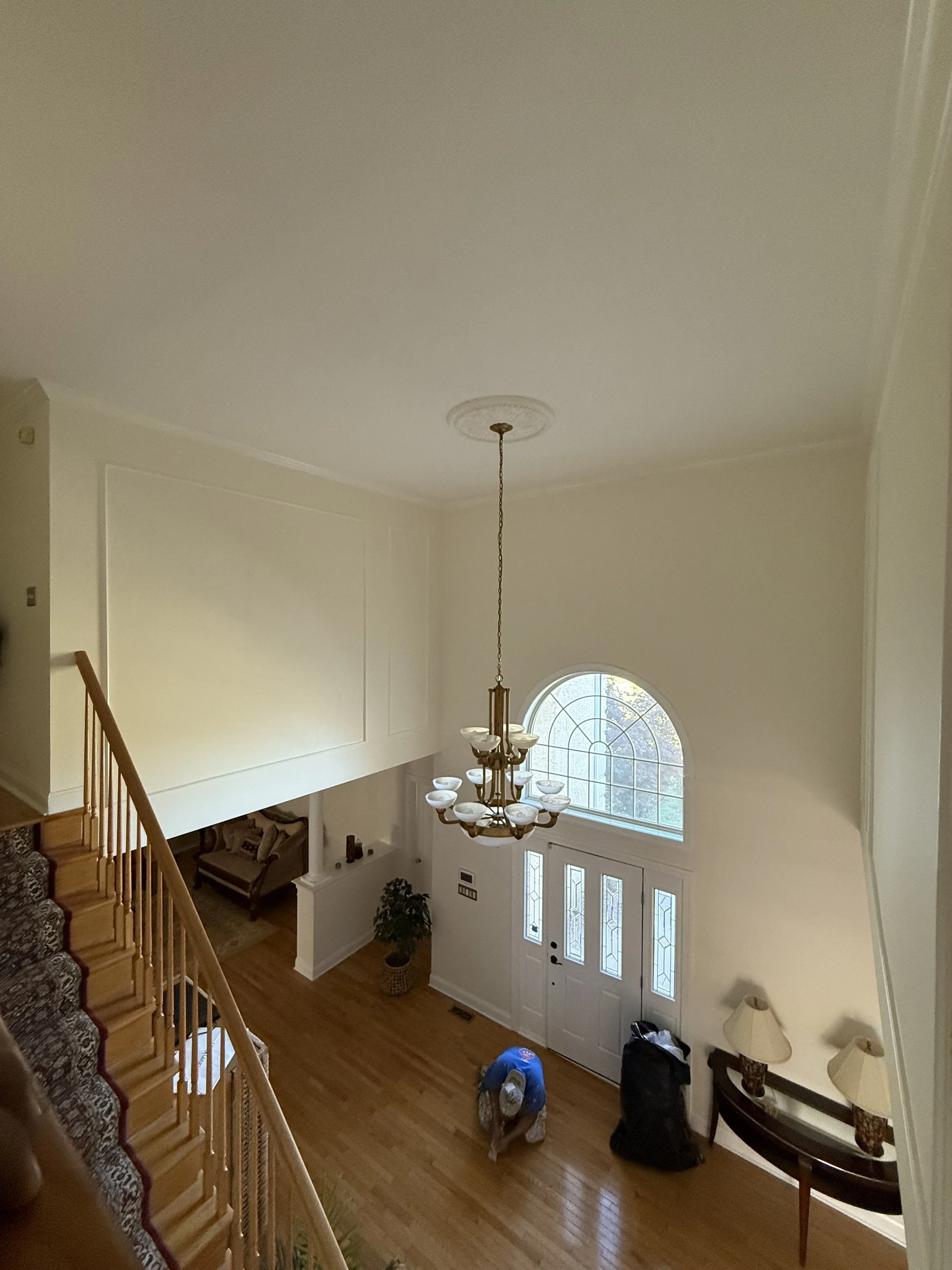 Top view of a spacious foyer with hardwood floors, a white front door with glass panels, a large arched window, a chandelier hanging from the ceiling, and a staircase with a patterned carpet on the left. There are a few bags, a potted plant, and two lamps on a side table near the entrance.