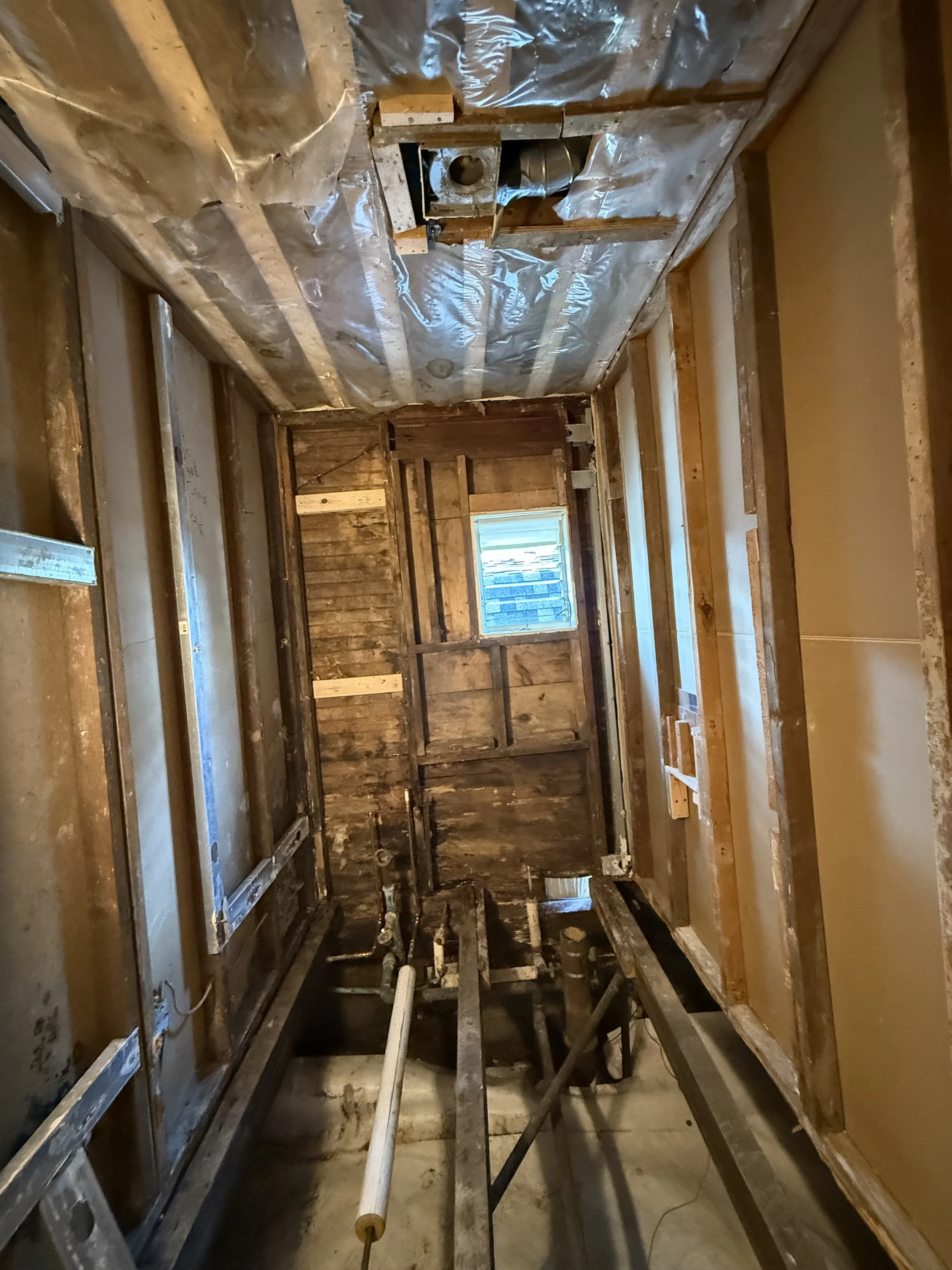 The interior of a room under renovation with exposed wooden framing, plumbing pipes, a small window, and construction materials.