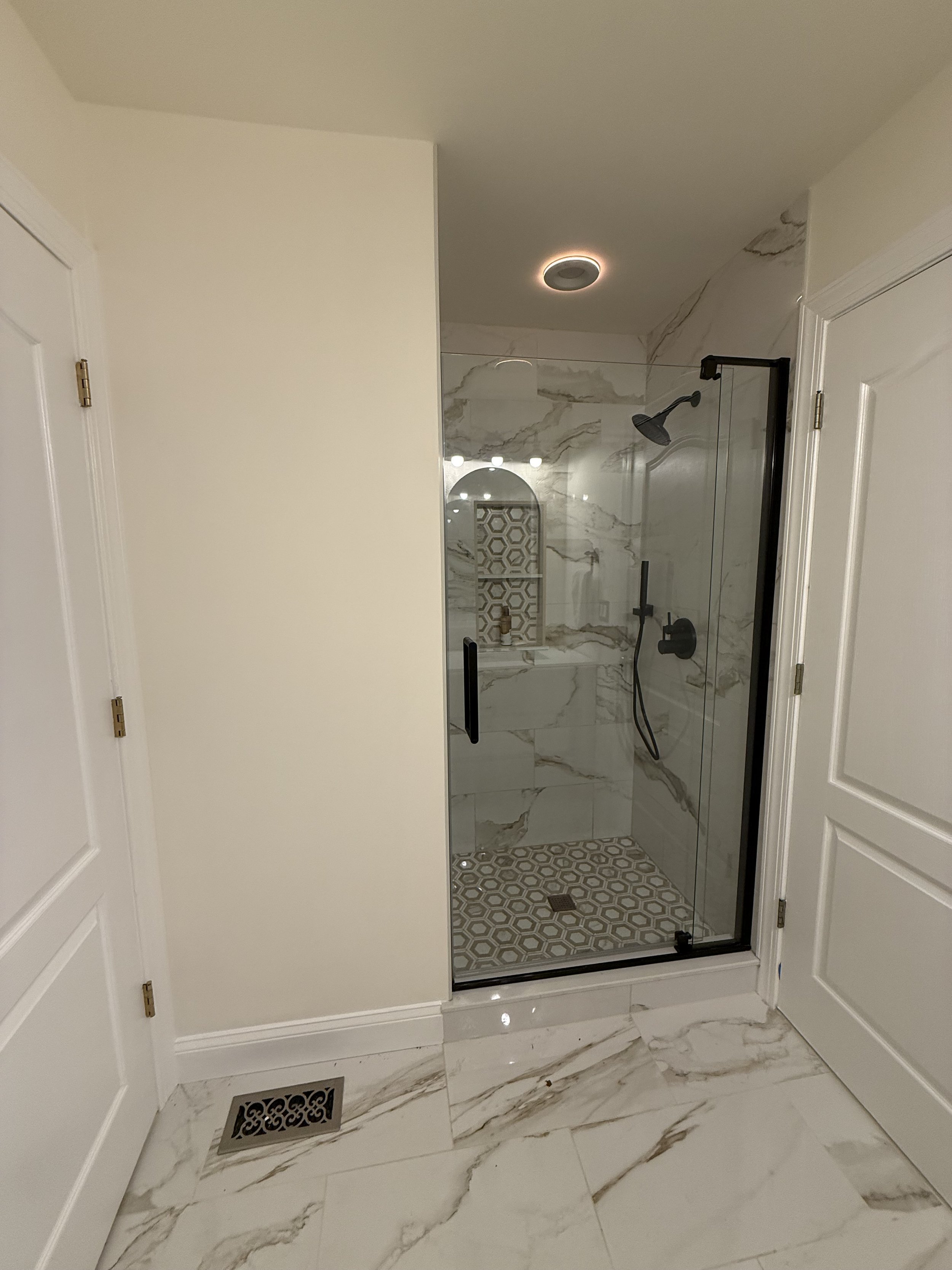 Modern walk-in shower with black fixtures, marble walls, sliding glass door, and patterned floor tiles, in a bathroom with marble flooring and white doors.