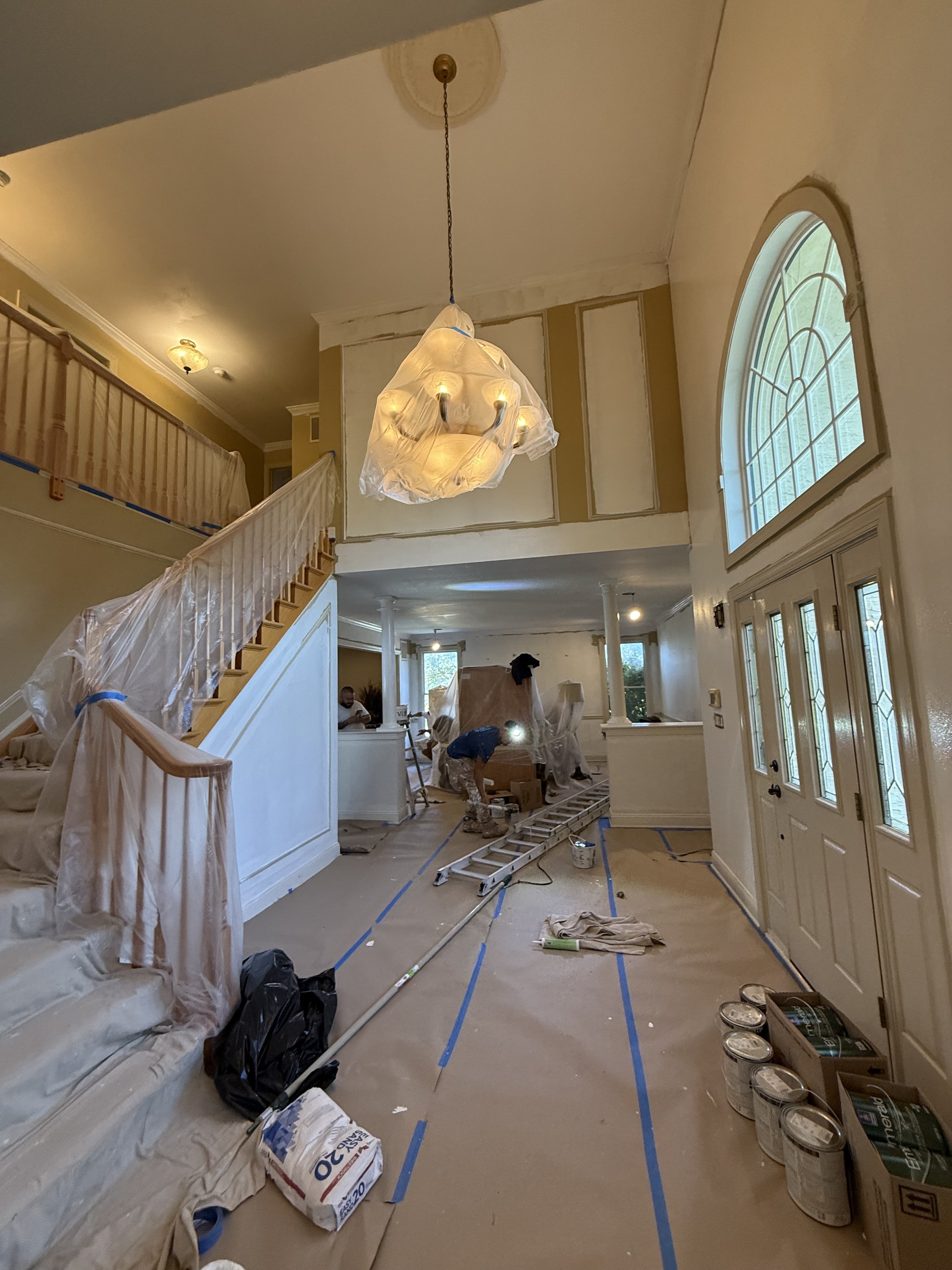 Living room under renovation with painters working, covered furniture, and renovation supplies on the floor.