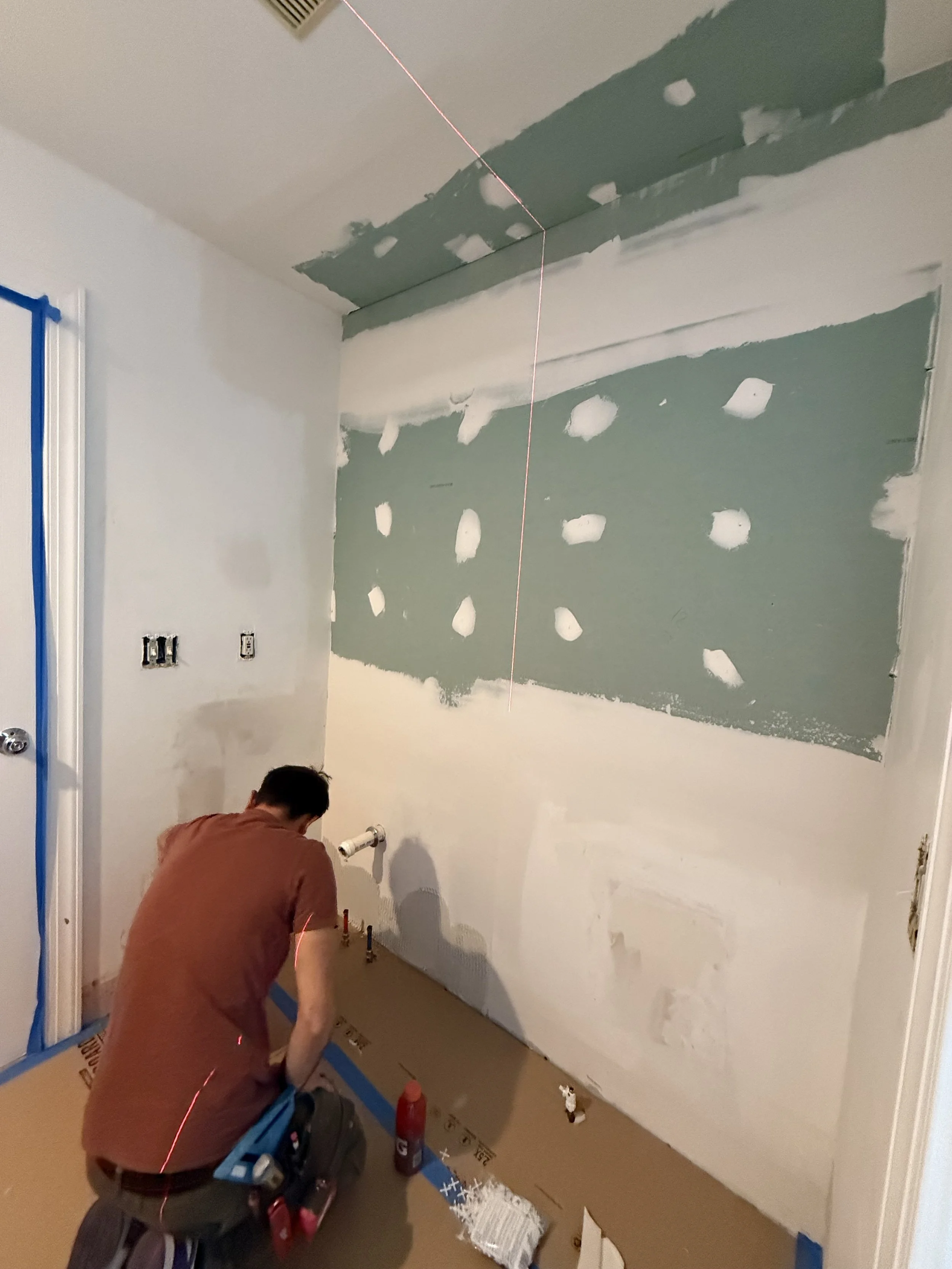 A person working on a drywall installation in a room, with a laser level projecting a red line across the ceiling and wall, and the wall partially painted with patches of drywall compound.