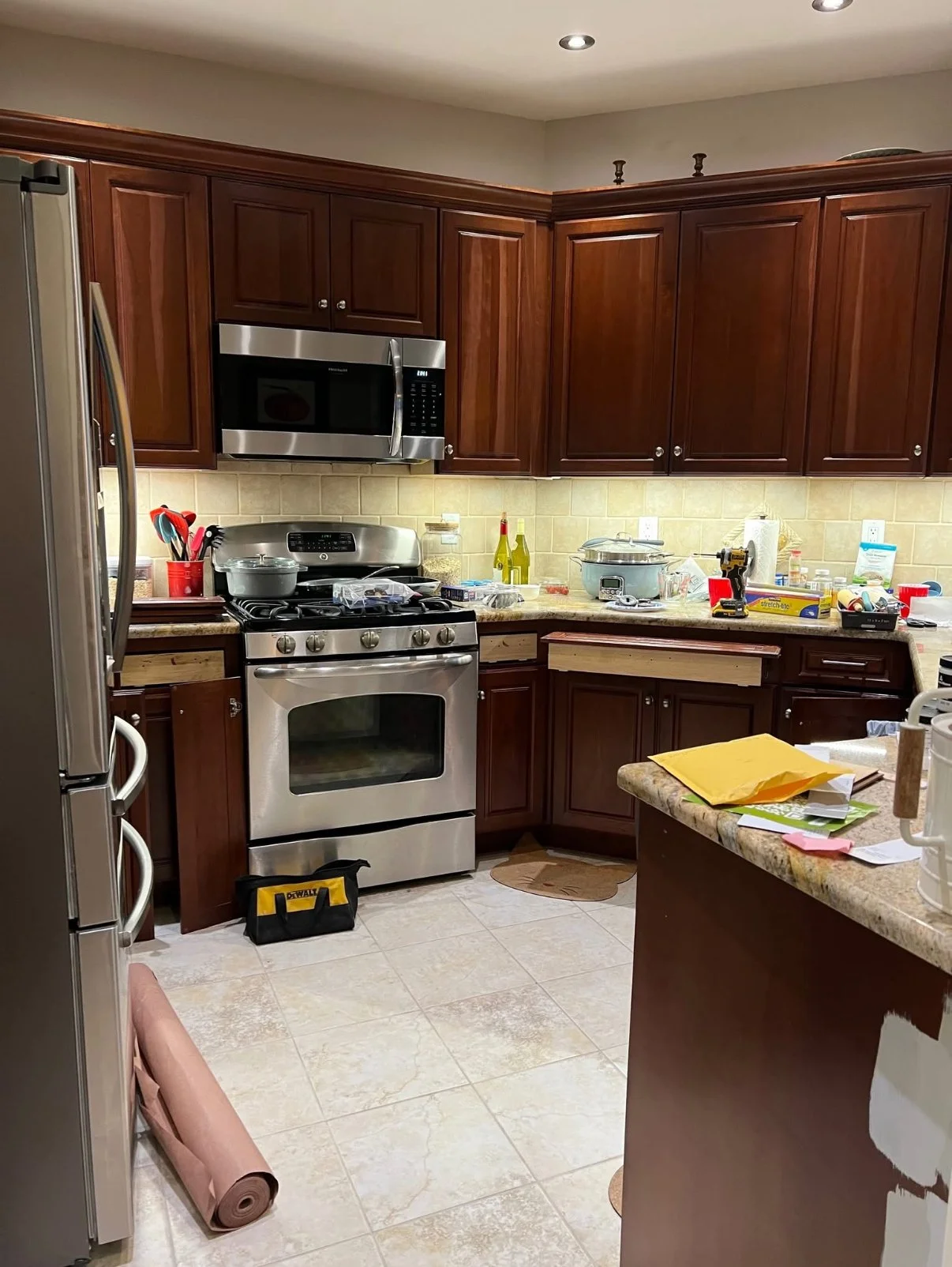 A cluttered kitchen with dark wooden cabinets, stainless steel appliances, and a granite countertop. The stove is in the center, with various items on the counter, including bottles, utensils, and containers. The floor has beige tiles, and there is a brown small mat near the stove.