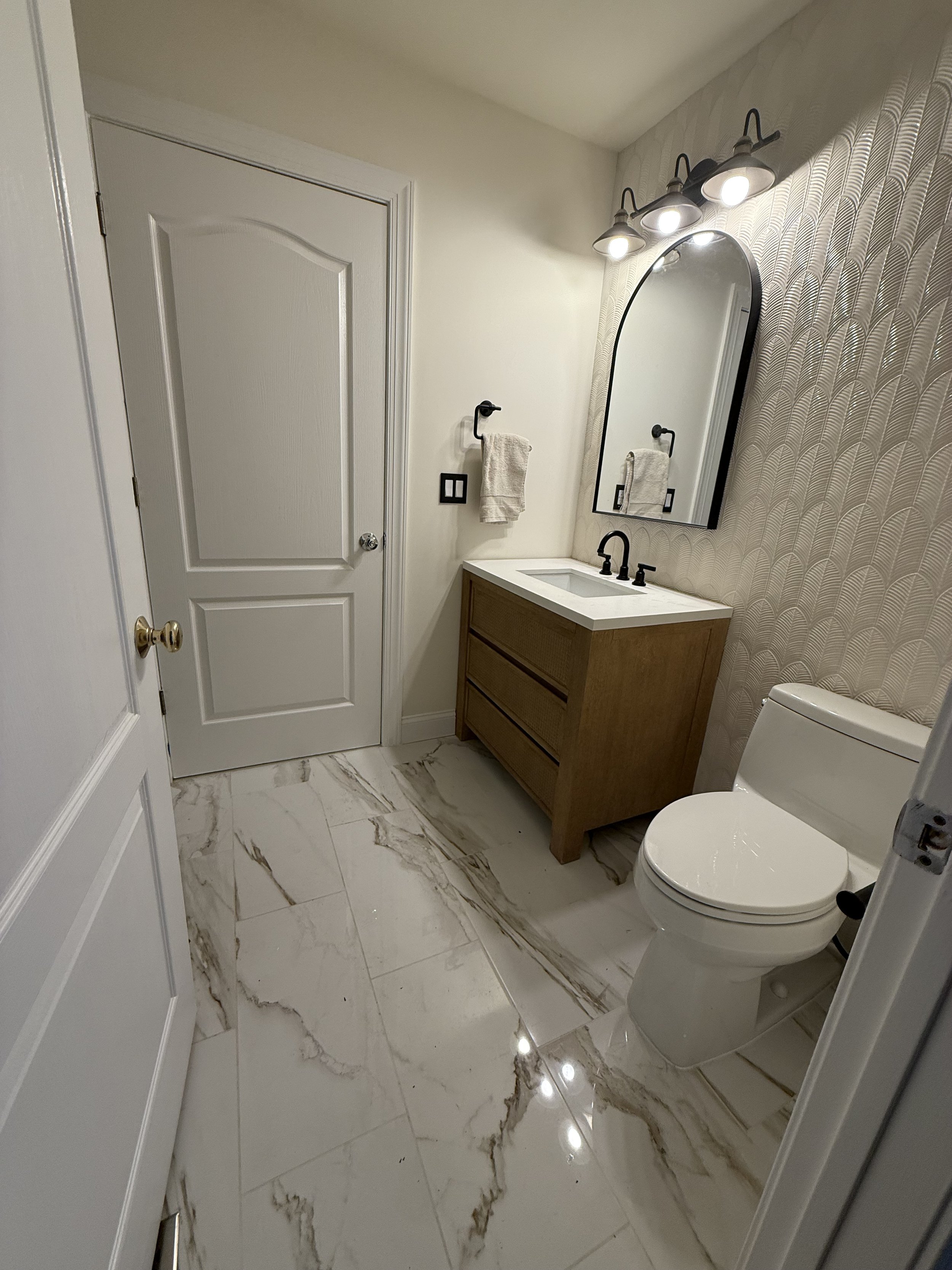 A modern bathroom with a marble floor, a white door, a wooden vanity with a white countertop and a black faucet, a round mirror, a three-light fixtures above the mirror, a towel hanging on a black towel holder, and a white toilet.