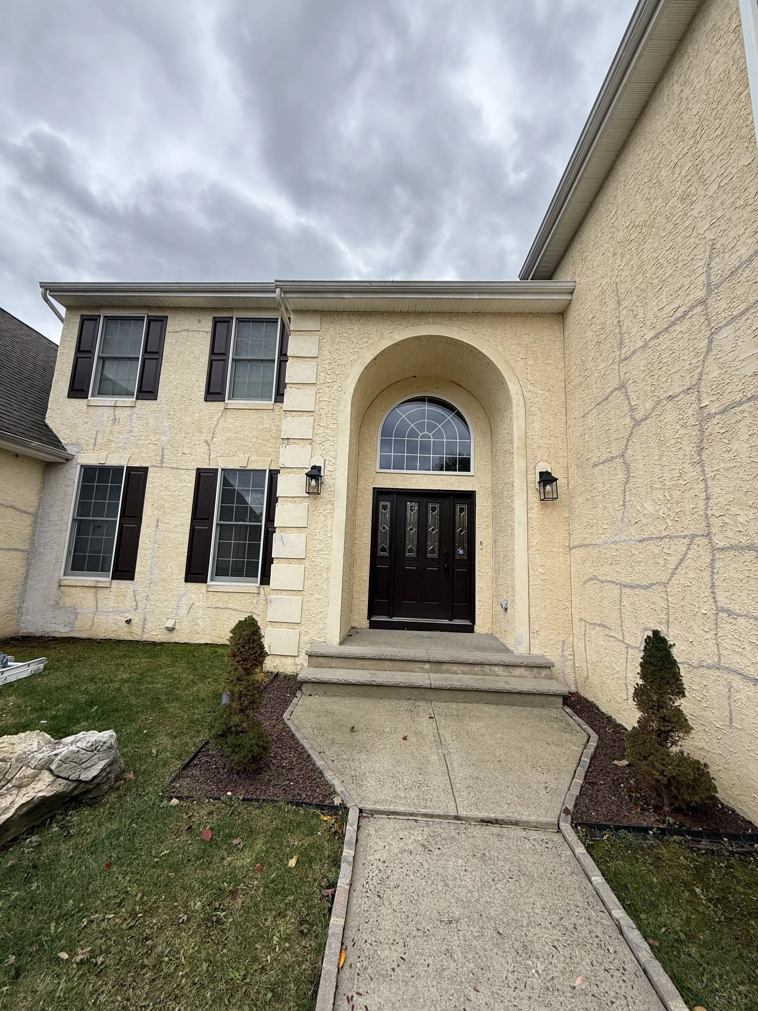 Front entrance of a yellow stucco house with a black front door, arched window above, and six windows with black shutters, two on each side of the door, under a cloudy sky.