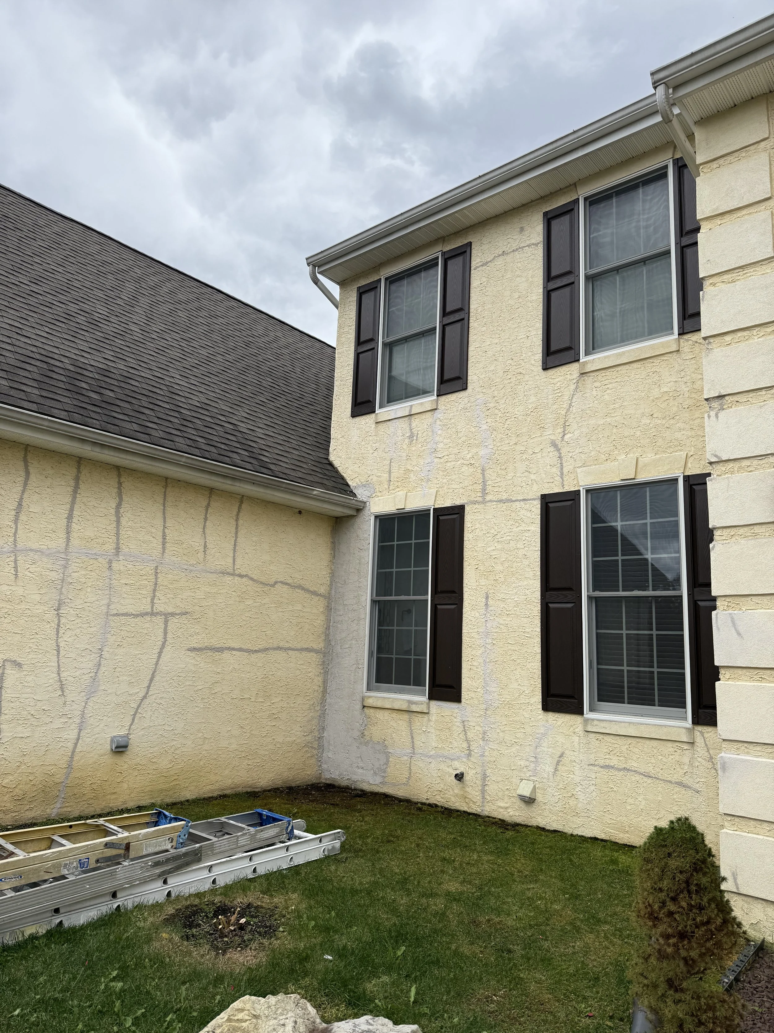 Backyard of a house with beige and yellow stucco walls, four windows with black shutters, a gray shingled roof, and a small shrub, with a ladder on the grass and a cloudy sky above.