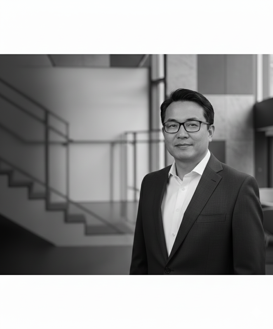 A black and white photograph of a man wearing glasses, a white shirt, and a dark suit, standing indoors with a staircase and modern architecture in the background.