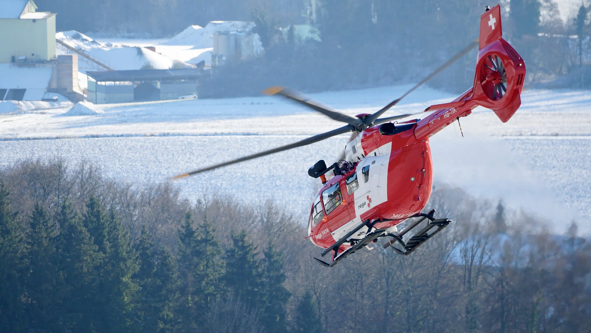 Rotes Rettungshelikopter fliegt in die Luft über eine winterliche Landschaft mit Schnee bedeckten Wiesen und Bäumen im Hintergrund.