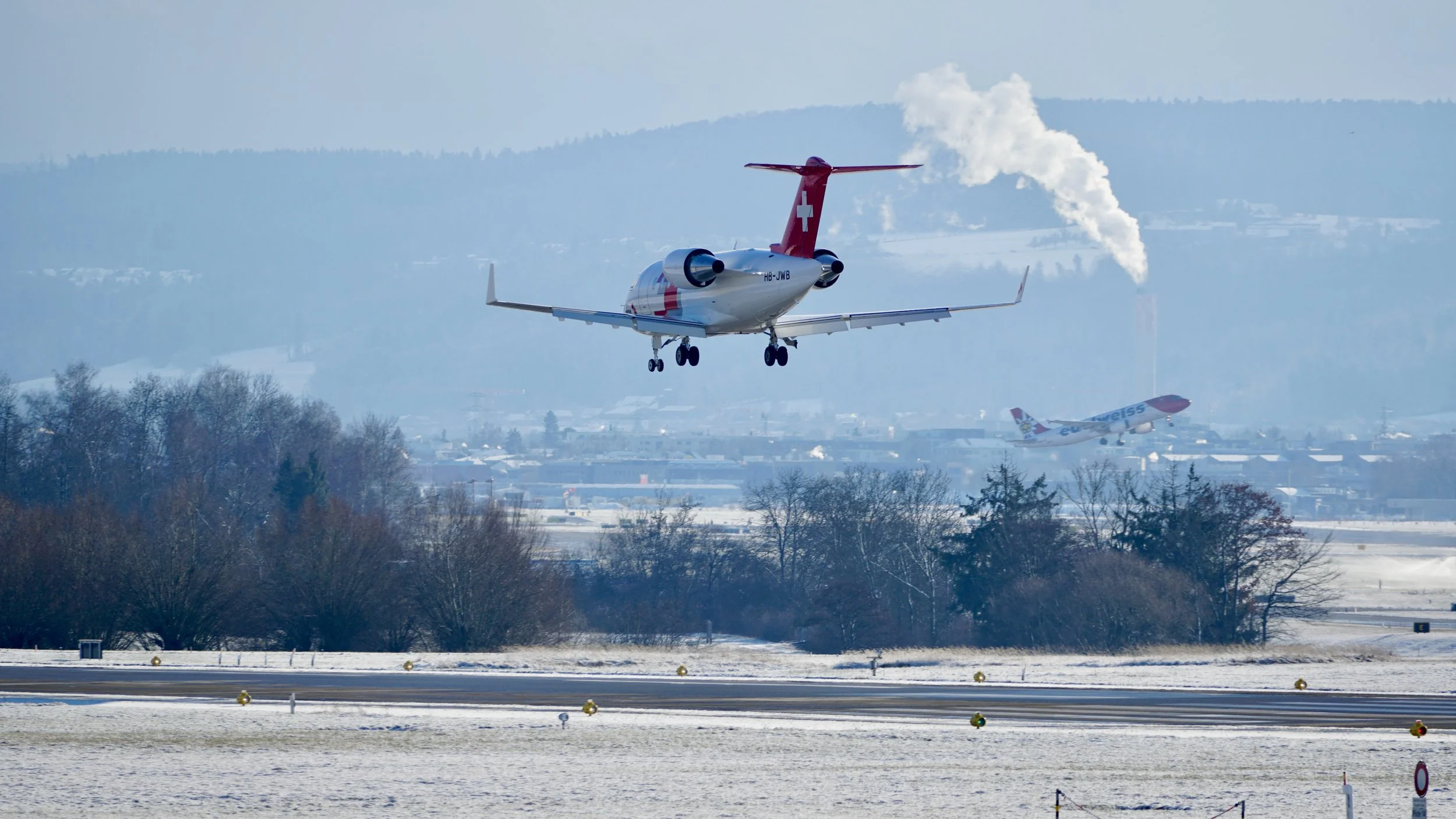 Ein Flugzeug fliegt über einen verschneiten Flughafen mit Bäumen und Hügel im Hintergrund, im Himmel sind weitere Flugzeuge zu sehen.
