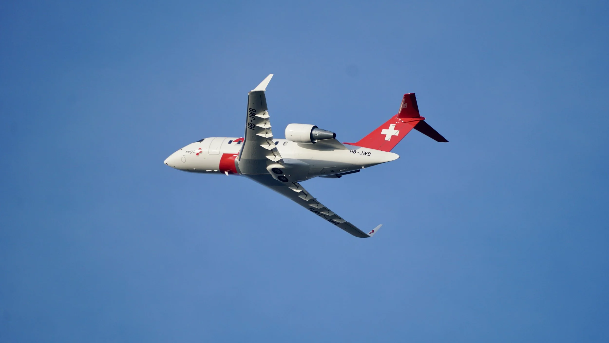 Ein weißes Flugzeug mit roten Akzenten und Schweizerfahne fliegt am blauen Himmel.