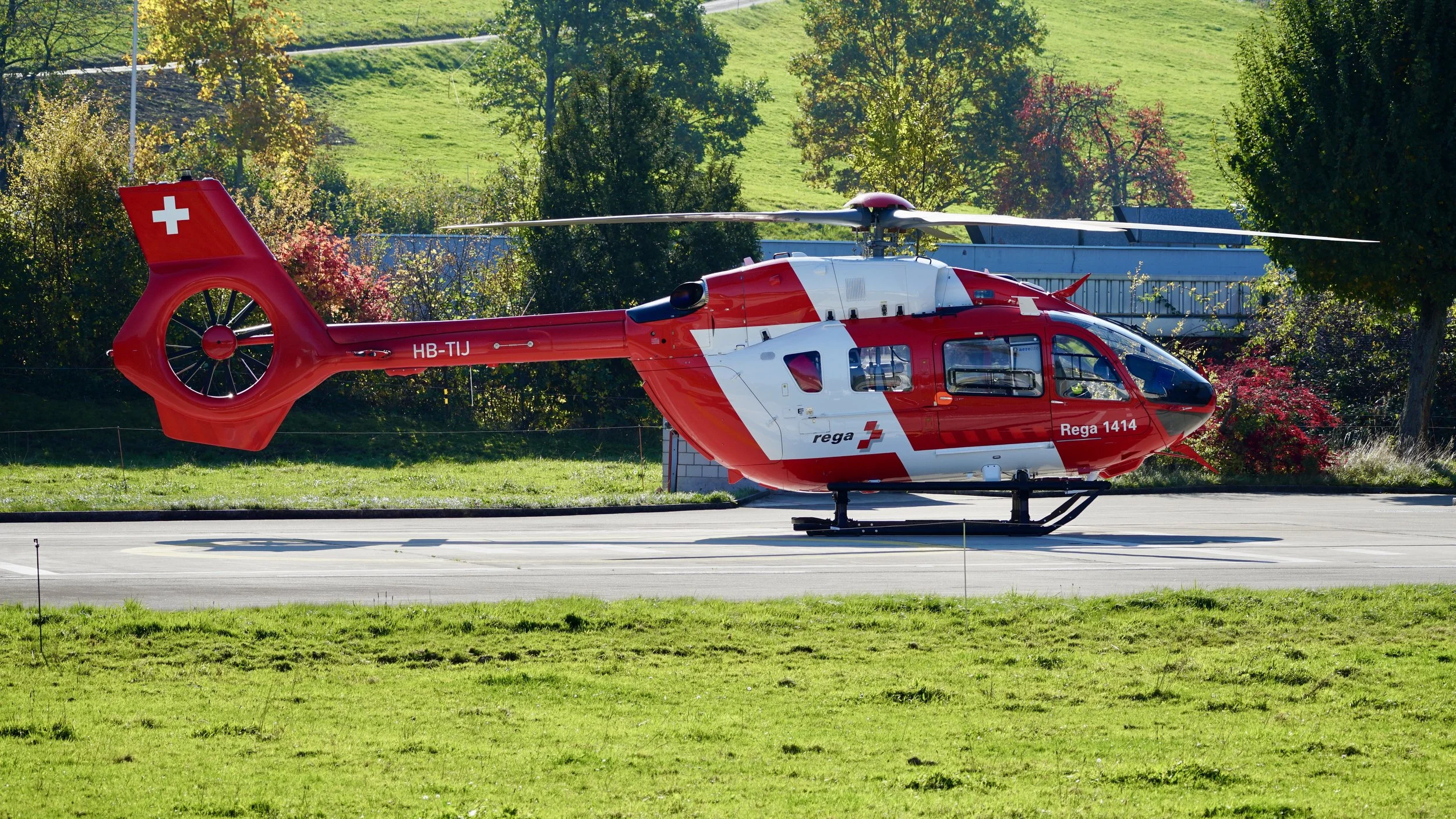 Rot-weisser Rettungshelikopter auf einer Flugpiste, im Hintergrund Bäume und grünes Gelände, mit Schweizer Flagge auf dem Heck