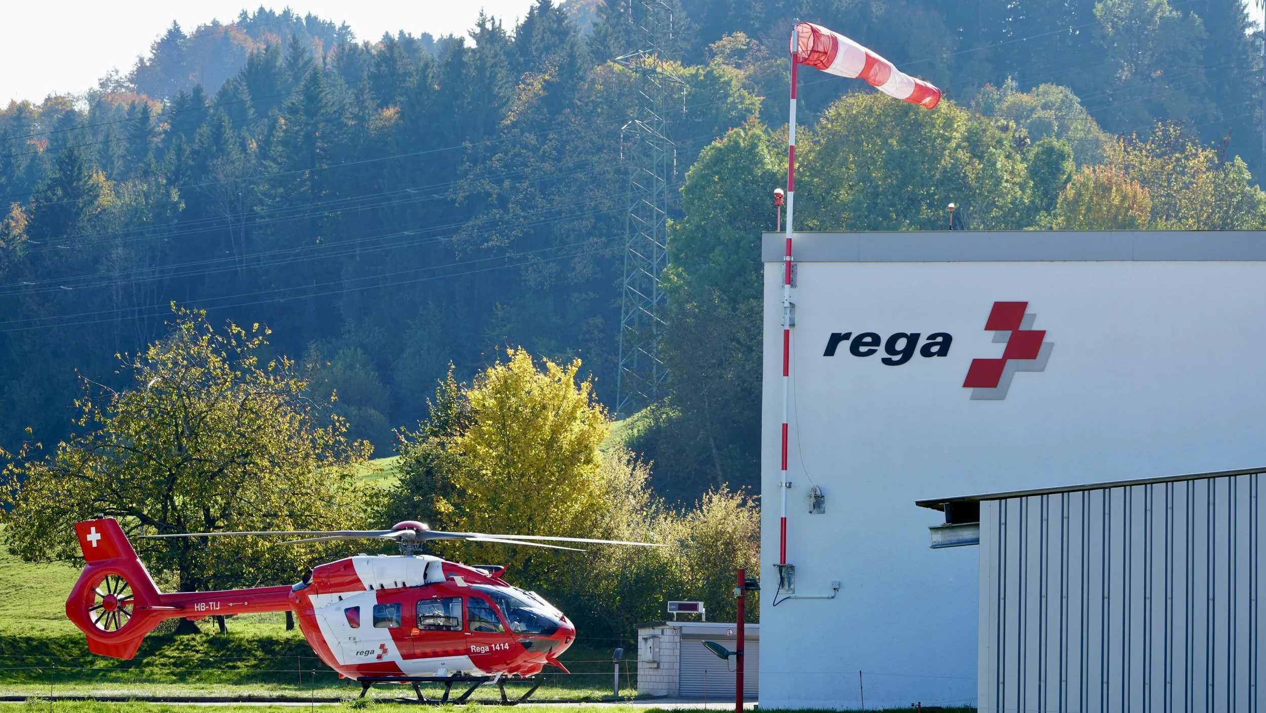 Roter Rettungshubschrauber der Rega steht auf dem Boden neben einem weißen Gebäude mit dem Rega-Logo, im Hintergrund Wälder und Berge.