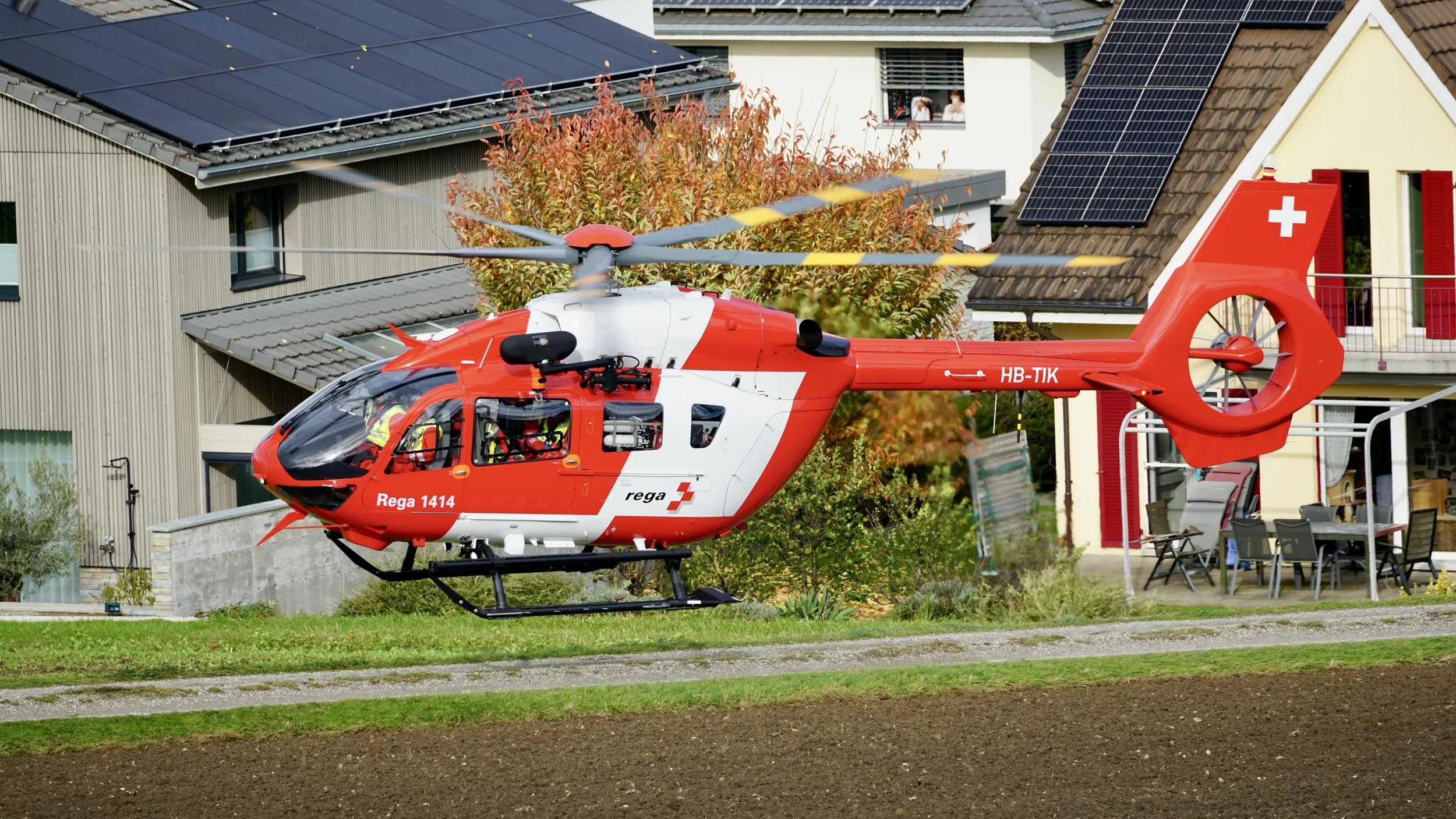Roter Rettungshelikopter schwebt über grünem Rasen vor Wohnhäusern mit Solardächern in einem Vorort.