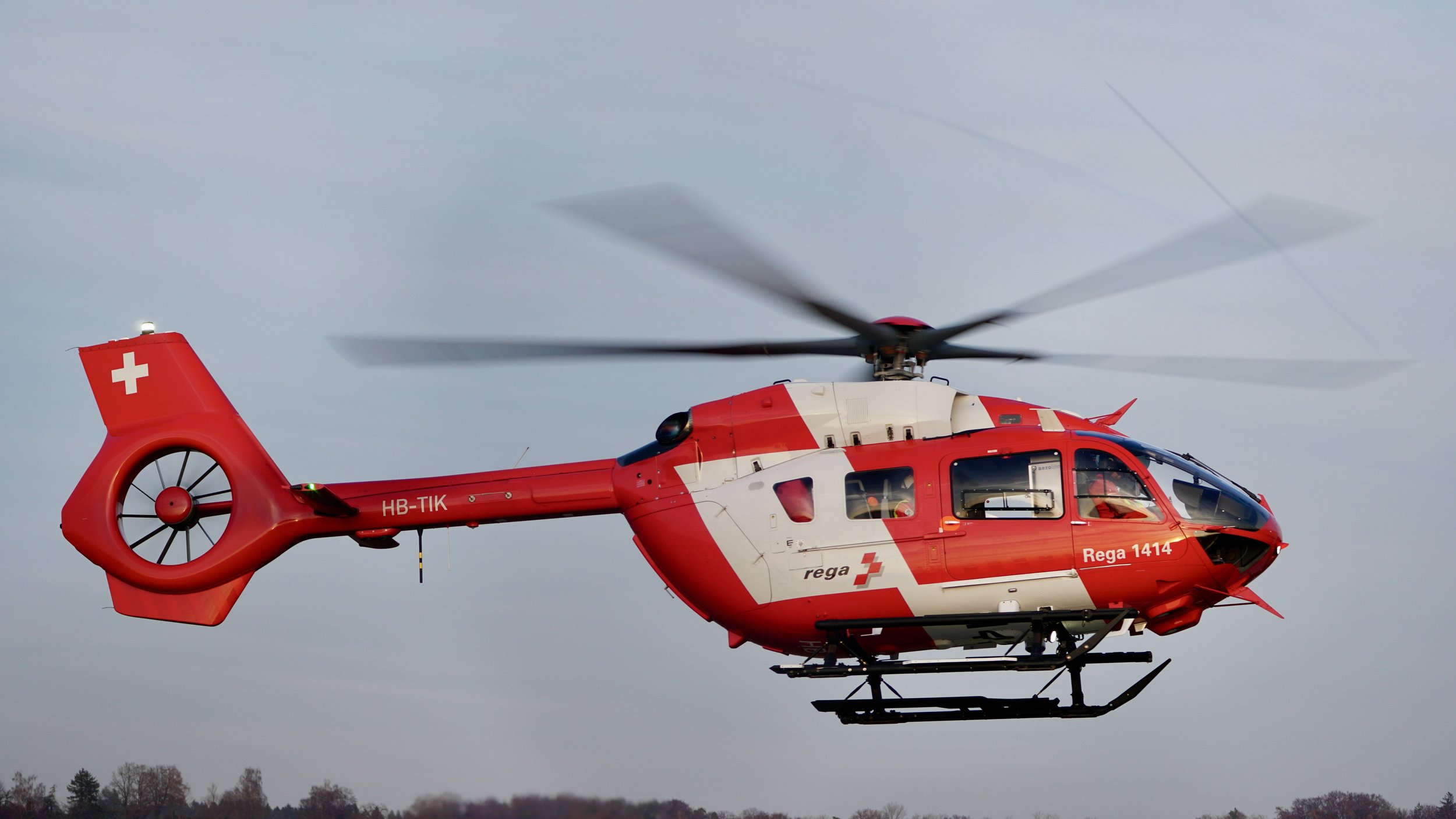 Ein roter Rettungshubschrauber fliegt über eine Landschaft mit Bäumen, ausgestattet mit weißen und roten Markierungen, inklusive einem weißen Kreuz auf dem Heck.