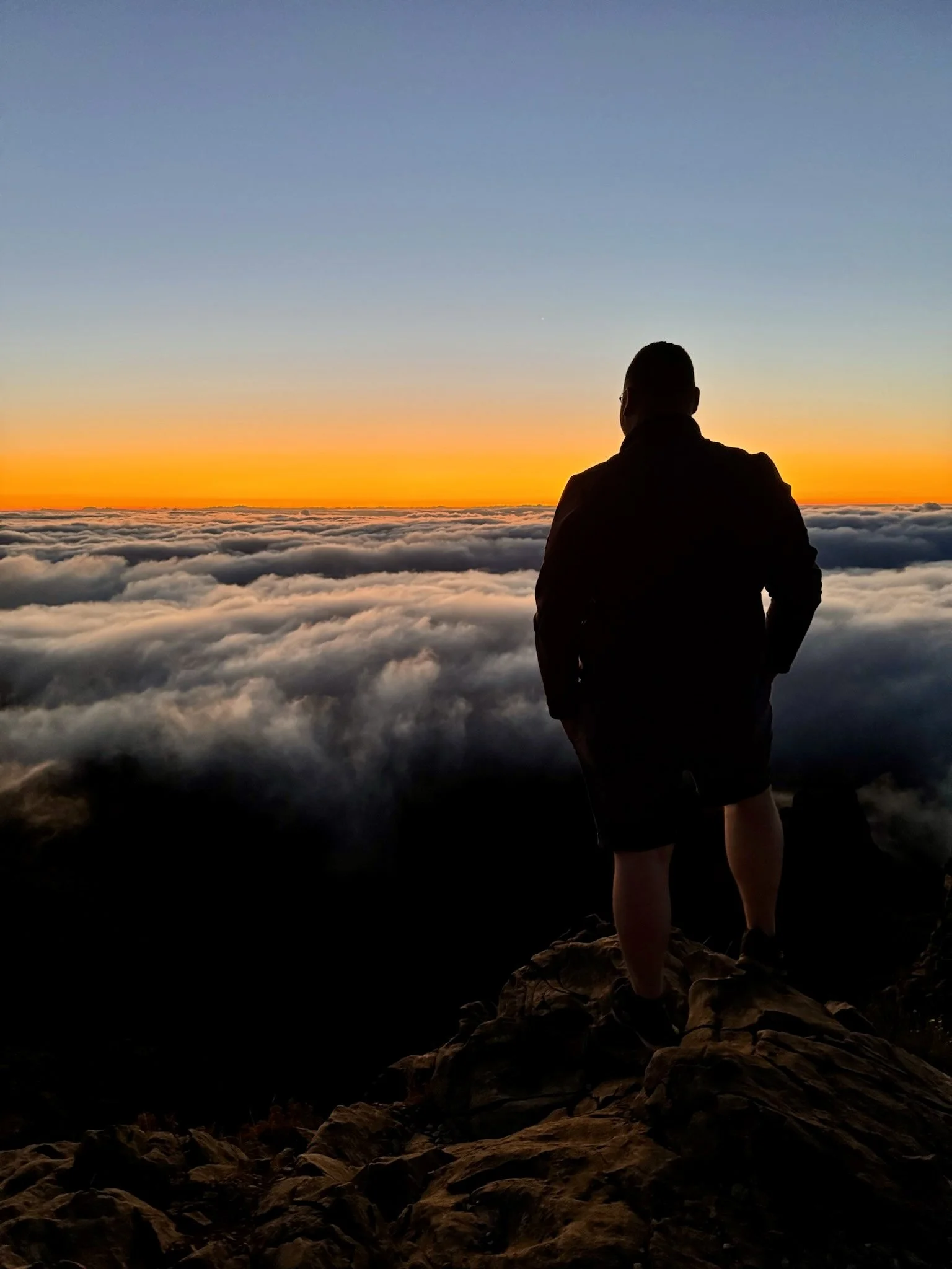 Eine Person steht auf einem Felsen und blickt auf eine Wolkendecke bei Sonnenaufgang, mit einem orangefarbenen Himmel im Hintergrund.