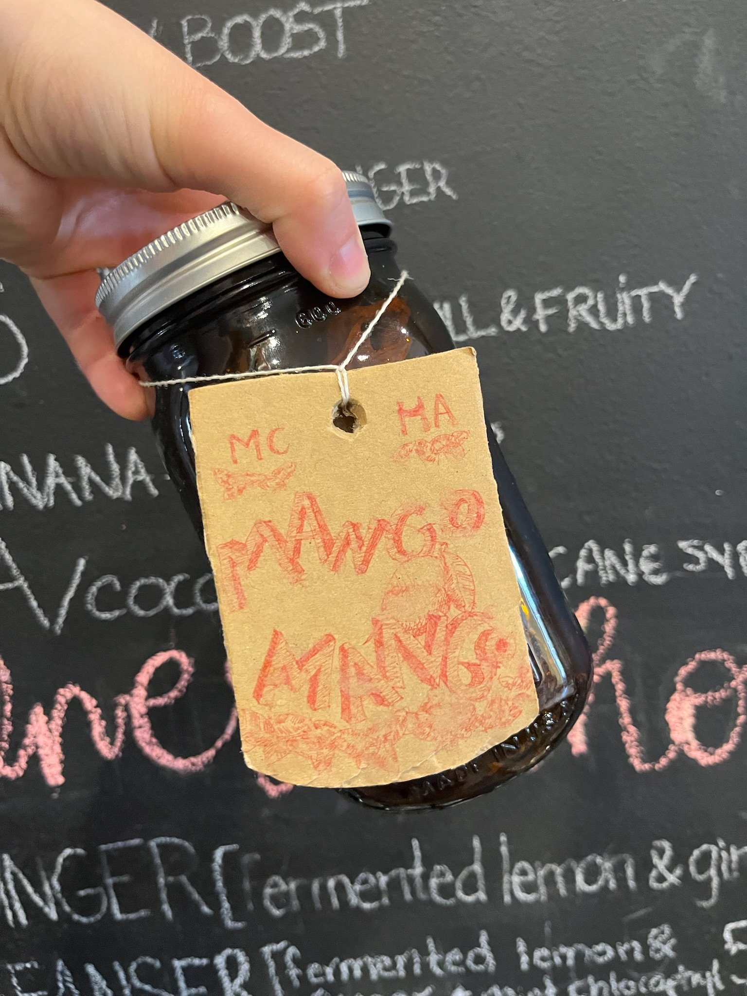 A hand holding a glass jar with a labeled tag hanging from it, in front of a blackboard with chalk writing.