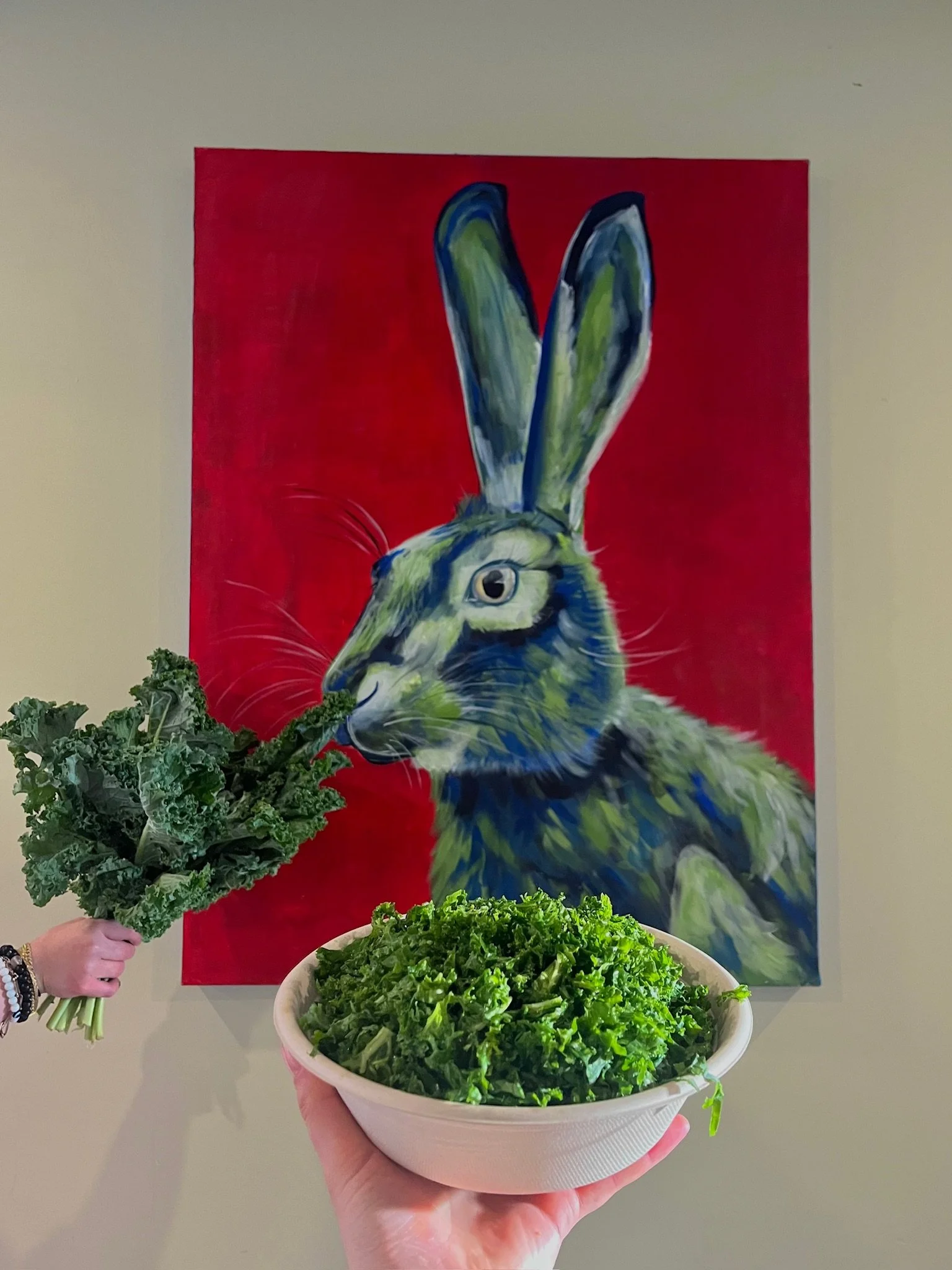 Person holding a bowl of kale in front of a colorful painting of a rabbit with kale in its mouth.
