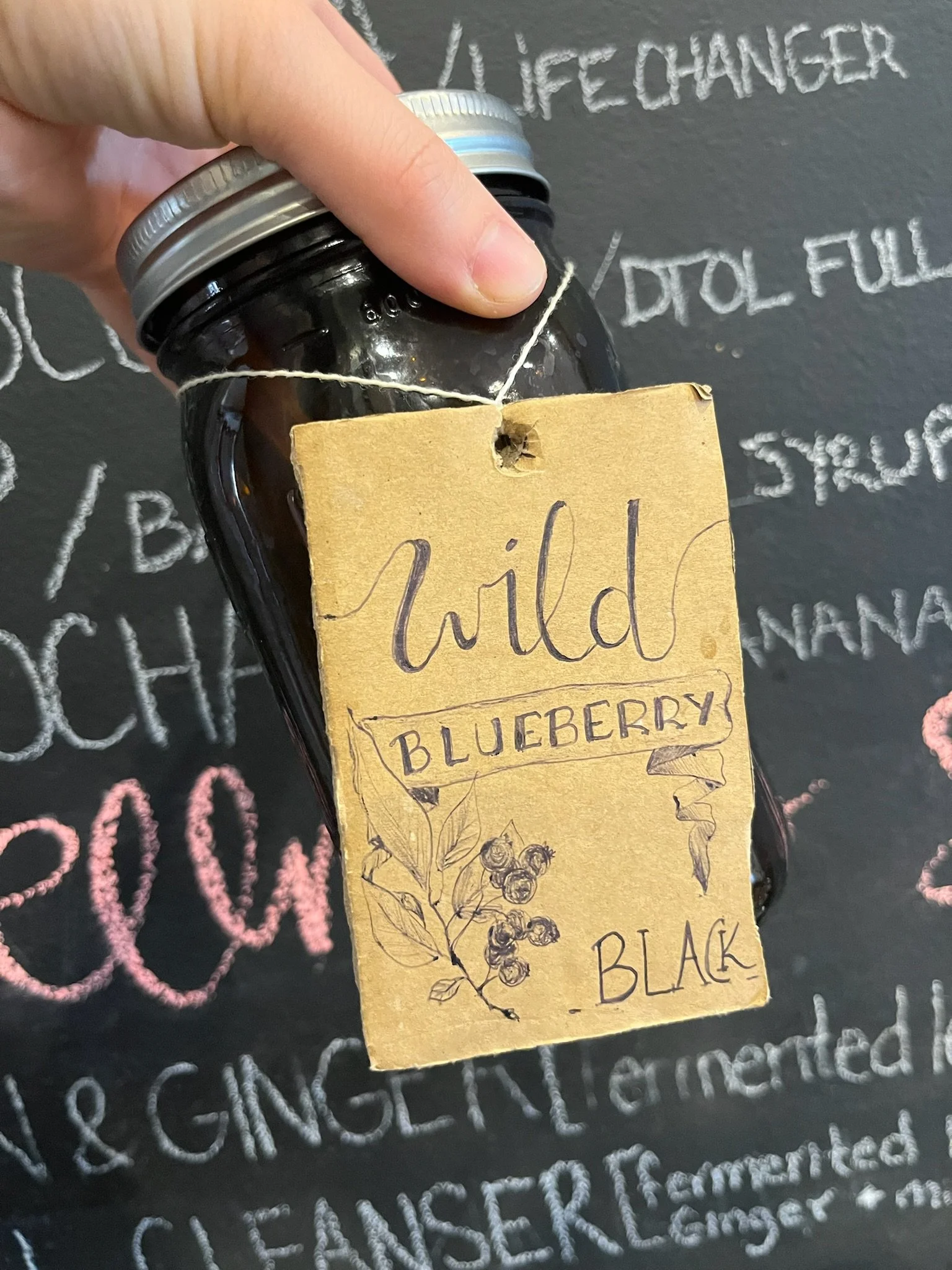 A hand holding a dark glass jar with a silver lid, attached to a brown paper label that reads "wild blueberry black" with an illustrated branch of blueberries. The background is a chalkboard with white chalk writing.