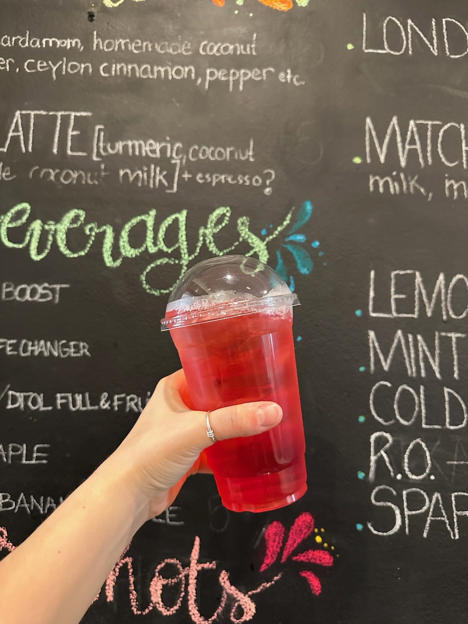 A person holding a clear red cold beverage in a plastic cup with a dome lid in front of a chalkboard menu with colorful writing.