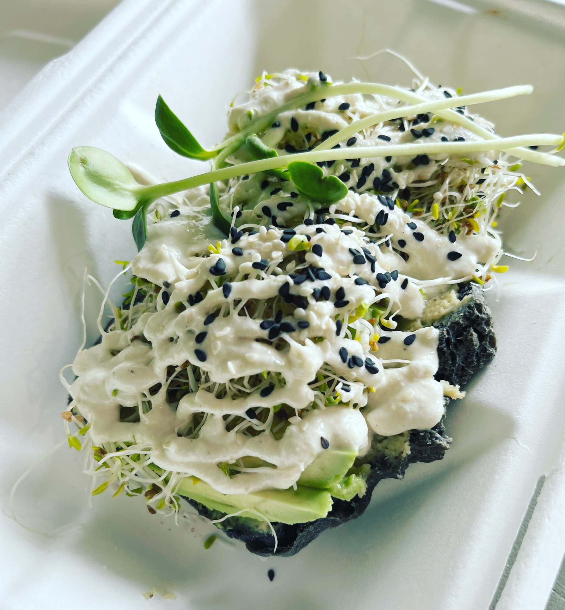 Close-up of a sushi roll topped with microgreens, black sesame seeds, and a creamy sauce, served on a white paper tray.