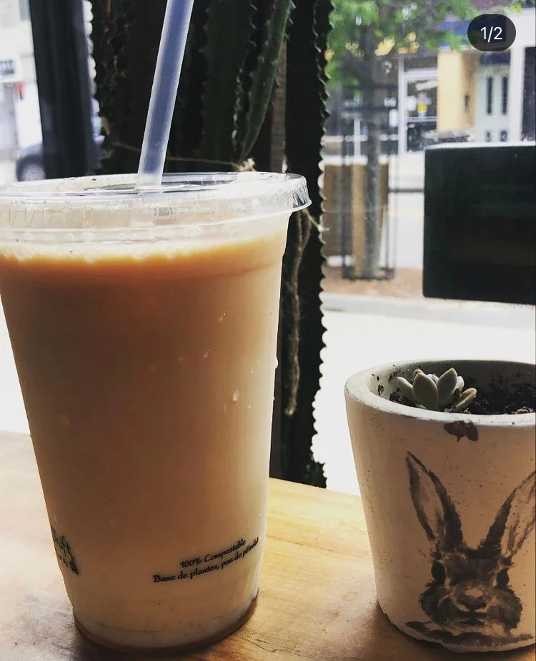 A plastic cup filled with iced coffee or latte with a straw, placed on a wooden table next to a small potted succulent plant with a bunny illustration on the pot, near a window with a street view outside.
